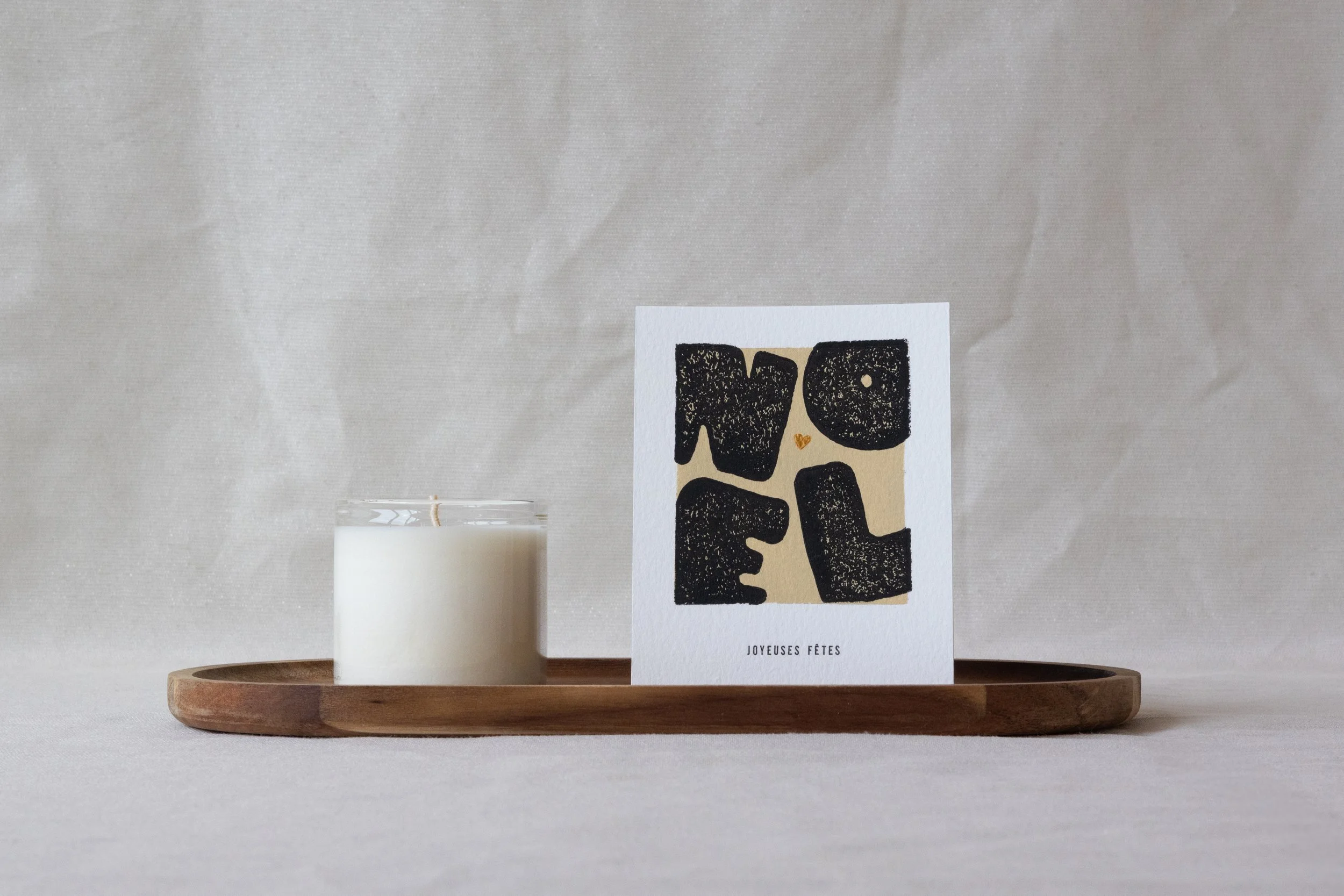 Une bougie blanche, une carte de vœux avec l'inscription 'NOEL' en lettres épaisses et noires, et le message 'JOYEUSES FÊTES' en bas, sur une planche en bois.