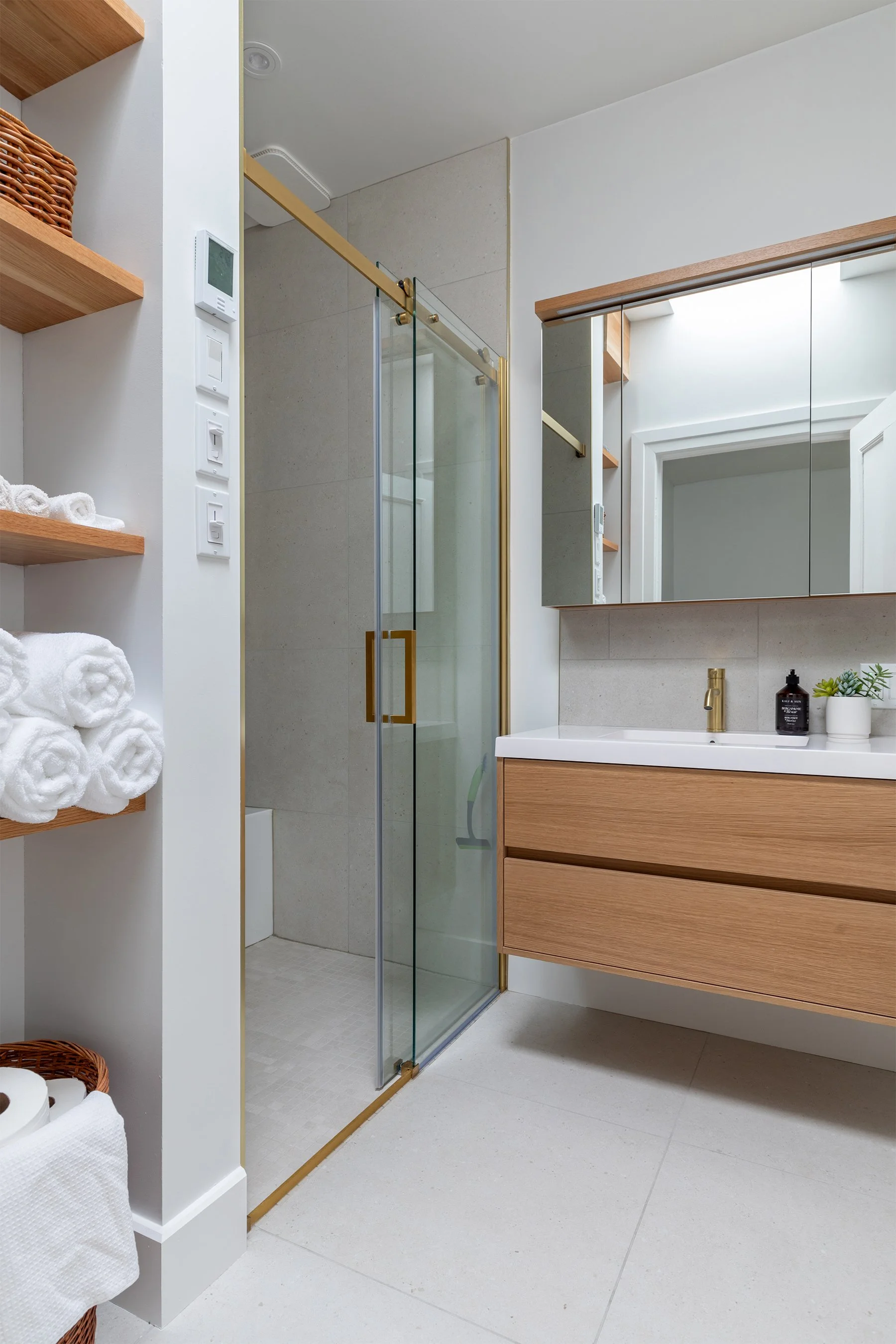 Salle de bain moderne avec cabine de douche en verre, meuble lavabo en bois et grand miroir, étagères avec serviettes blanches, et décor minimaliste.