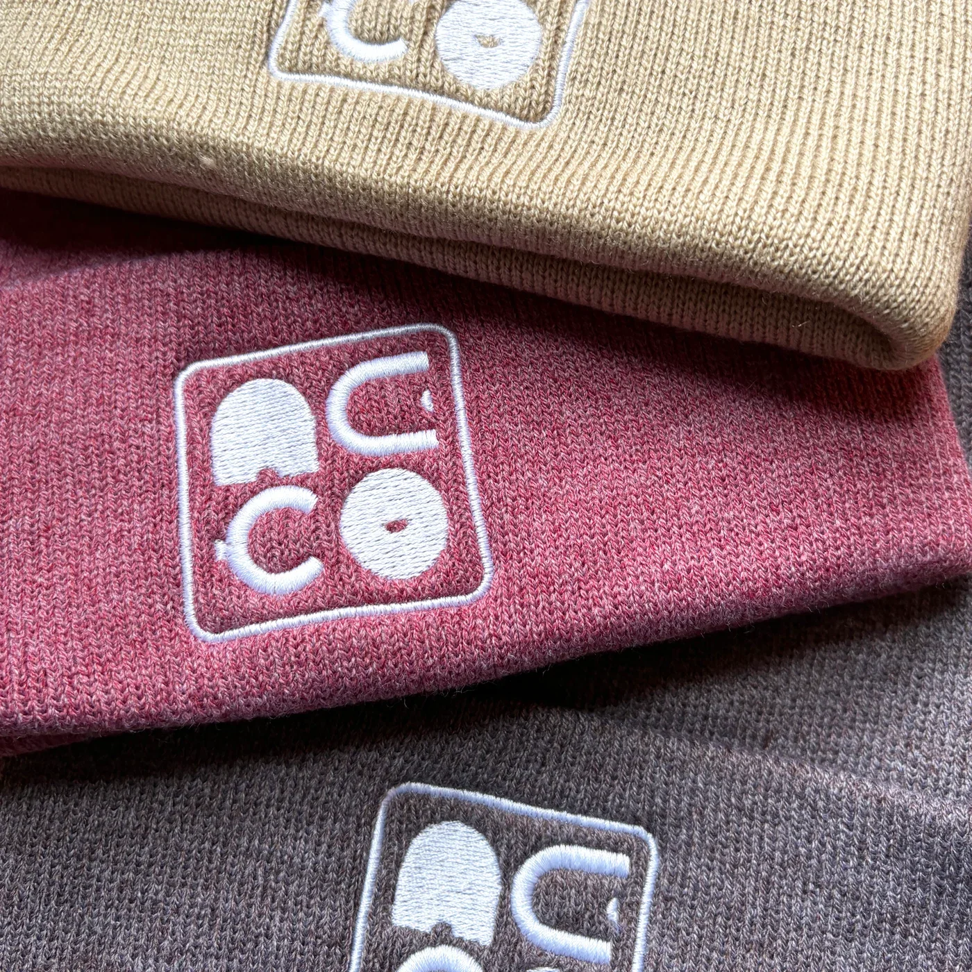 Trois bonnets en laine avec un logo brodé blanc, de couleur beige, rose et gris.