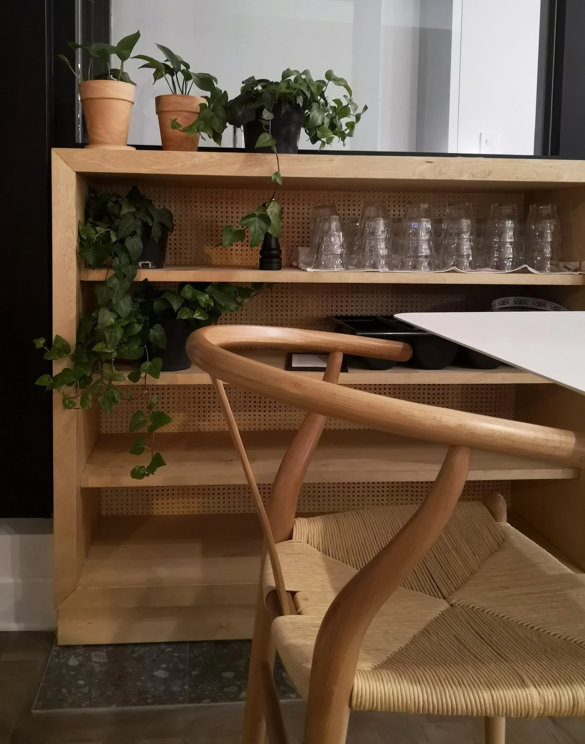 Une étagère en bois contenant des plantes en pot, des verres en verre empilés et des ustensiles noirs, avec un fauteuil en bois et une table blanche à l'avant.