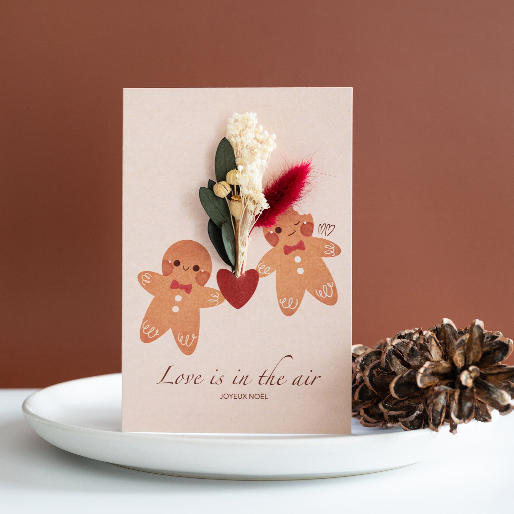 Carte de vœux de Noël avec deux bonhommes en pain d'épice, un cœur rouge, et des fleurs séchées attachées, le tout est disposé sur un plateau blanc orné d'une branche de pin.