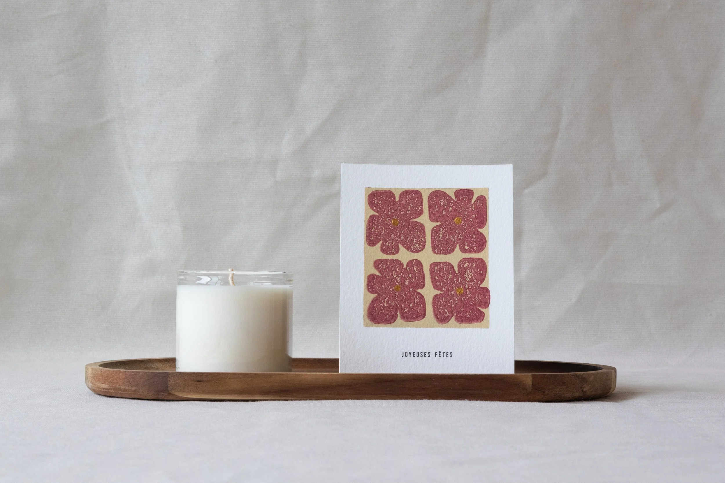 Carte de vœux avec un motif de fleurs roses imprimées avec la technique de la linogravure. La carte est posée au côté d'une bougie sur un plateau en bois avec un arrière plan beige.