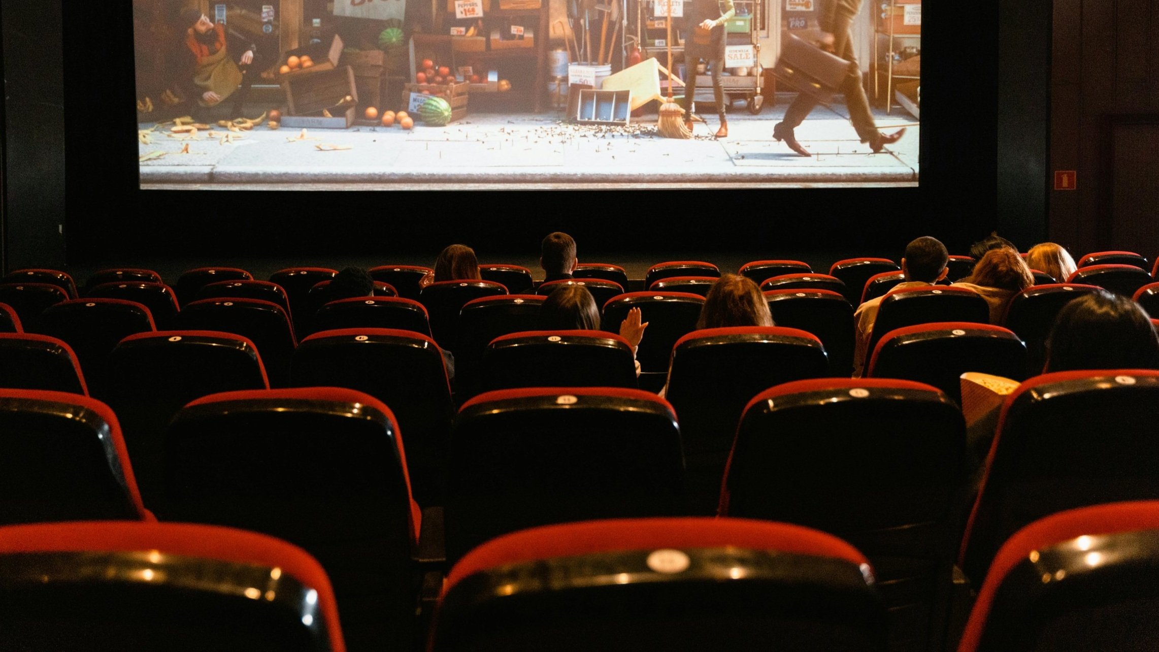 A movie theater scene facing the screen, with a movie shown in the background and people filling the seats in the foreground.