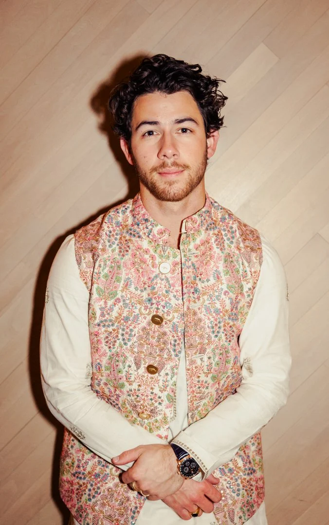 A man with dark, curly hair and a beard, wearing a cream-colored shirt with an ornate, pinkish floral embroidered vest, standing against a sparsely wooden backdrop.