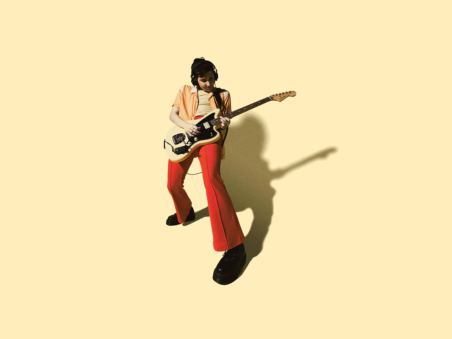Person wearing headphones playing an electric guitar on a yellow background, casting a shadow.