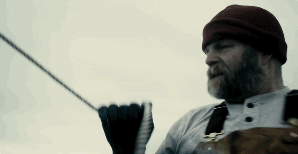 Man with a beard, wearing a maroon knit cap, gray shirt, and black glove holding a rope, outdoors with cloudy sky.