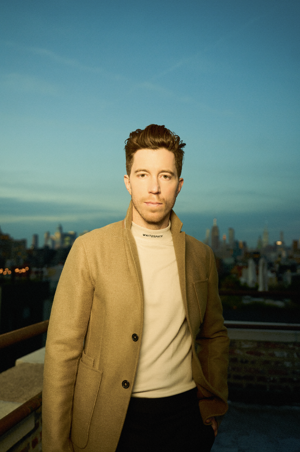 A young man with brown hair styled upwards, wearing a beige coat over a white shirt, standing outdoors during sunset with a city skyline in the background.