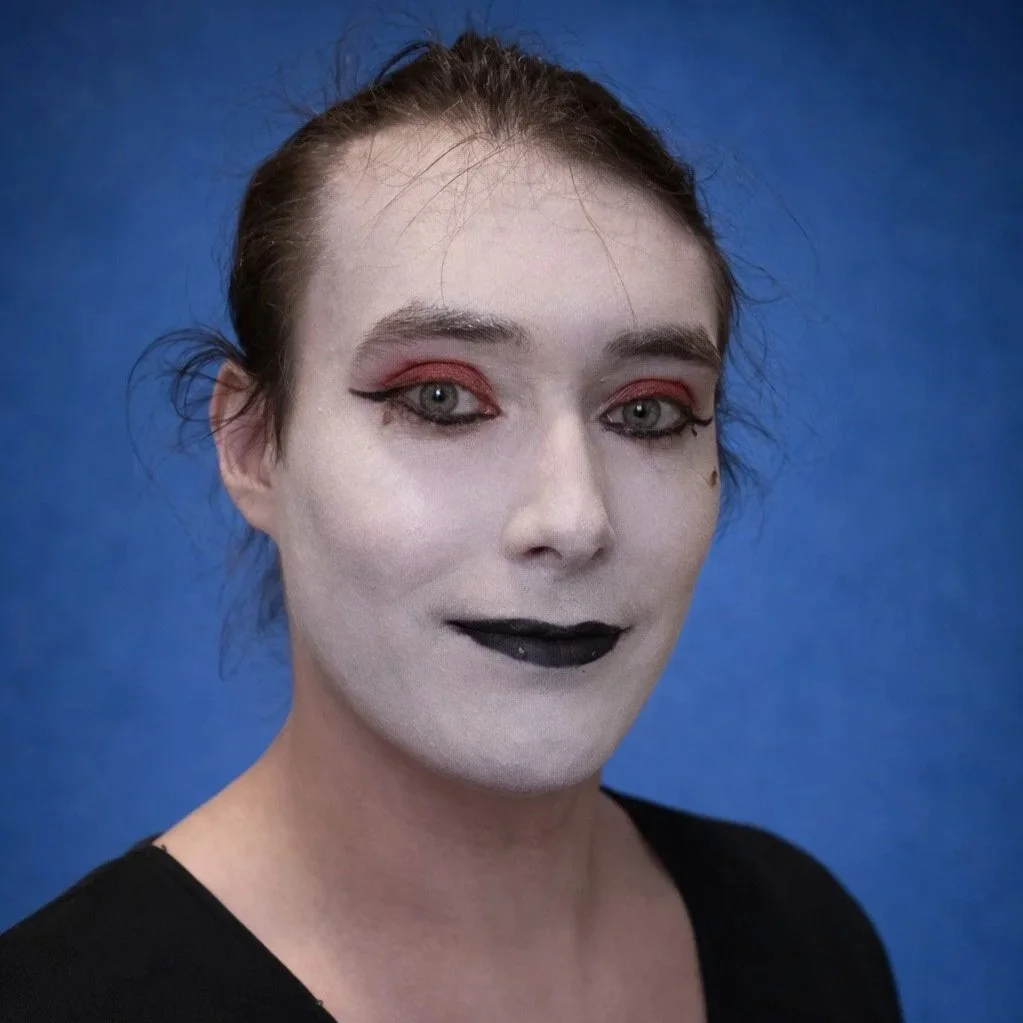 Heccatomb is a white transfeminine person with light skin and dark brown hair pulled back.  Her eyebrows are dark and defined, and her eyes are light colored. She is wearing a simple black V-neck top and stands against a solid blue background.