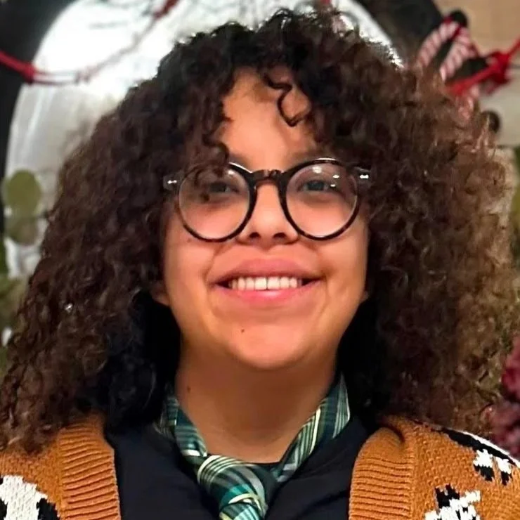 Alondra is a lesbian hispanic person with medium long curly hair. Alondra is wearing this round glasses with brown camo rim and a brown sweater with cows on it. She is wearing a green tie with a black shirt, smiling widely.