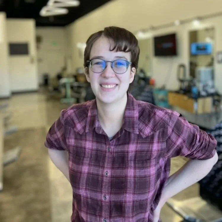 Ariana is a white genderqueer person with short brown hair and curly bangs. Ariana is wearing dark grey glasses with large round lenses. She has a pink plaid button-up shirt on and is smiling joyfully.