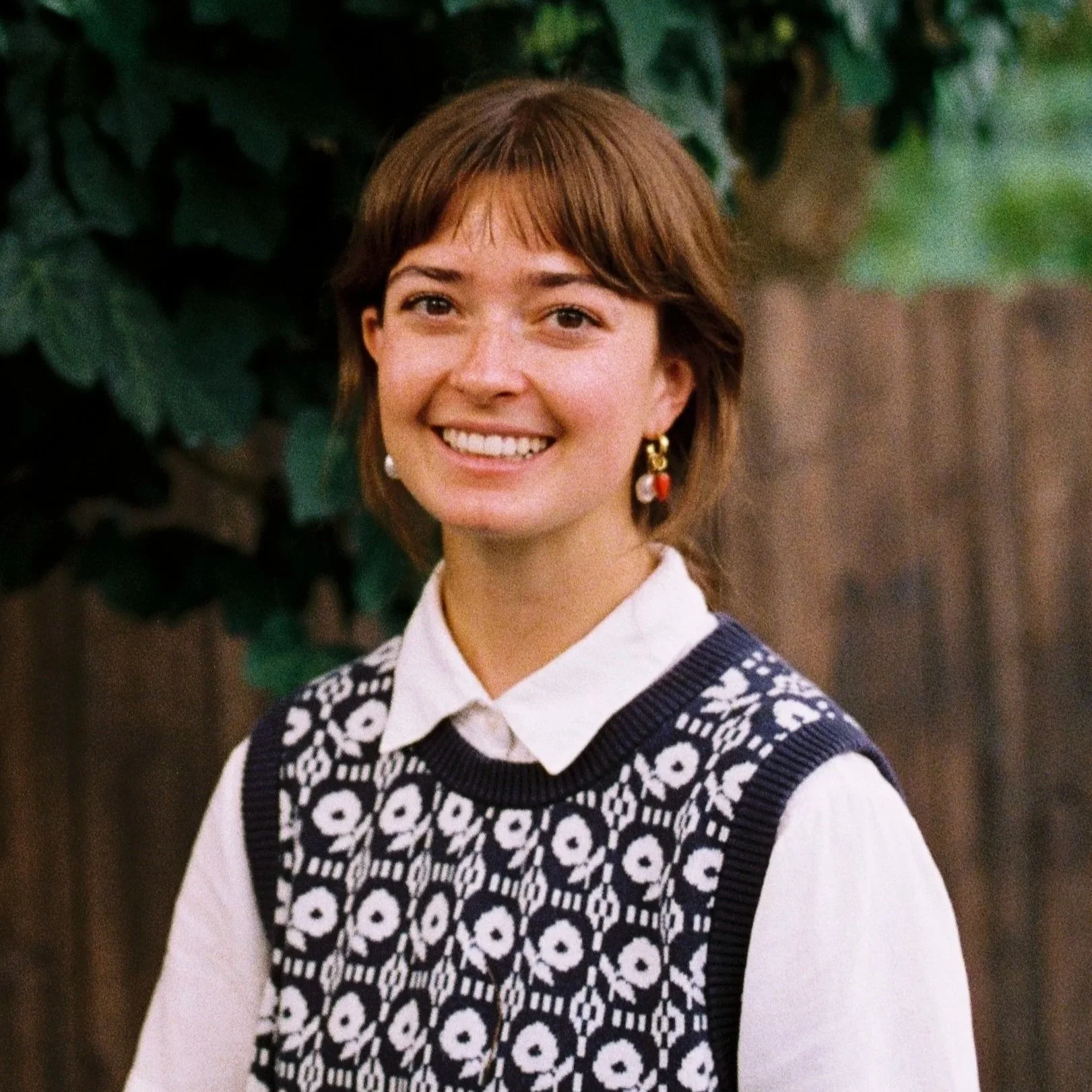 Paisley is a non-binary white person with short brown hair and bangs. They are wearing a navy blue, floral patterned sweater vest. The background is ivy.