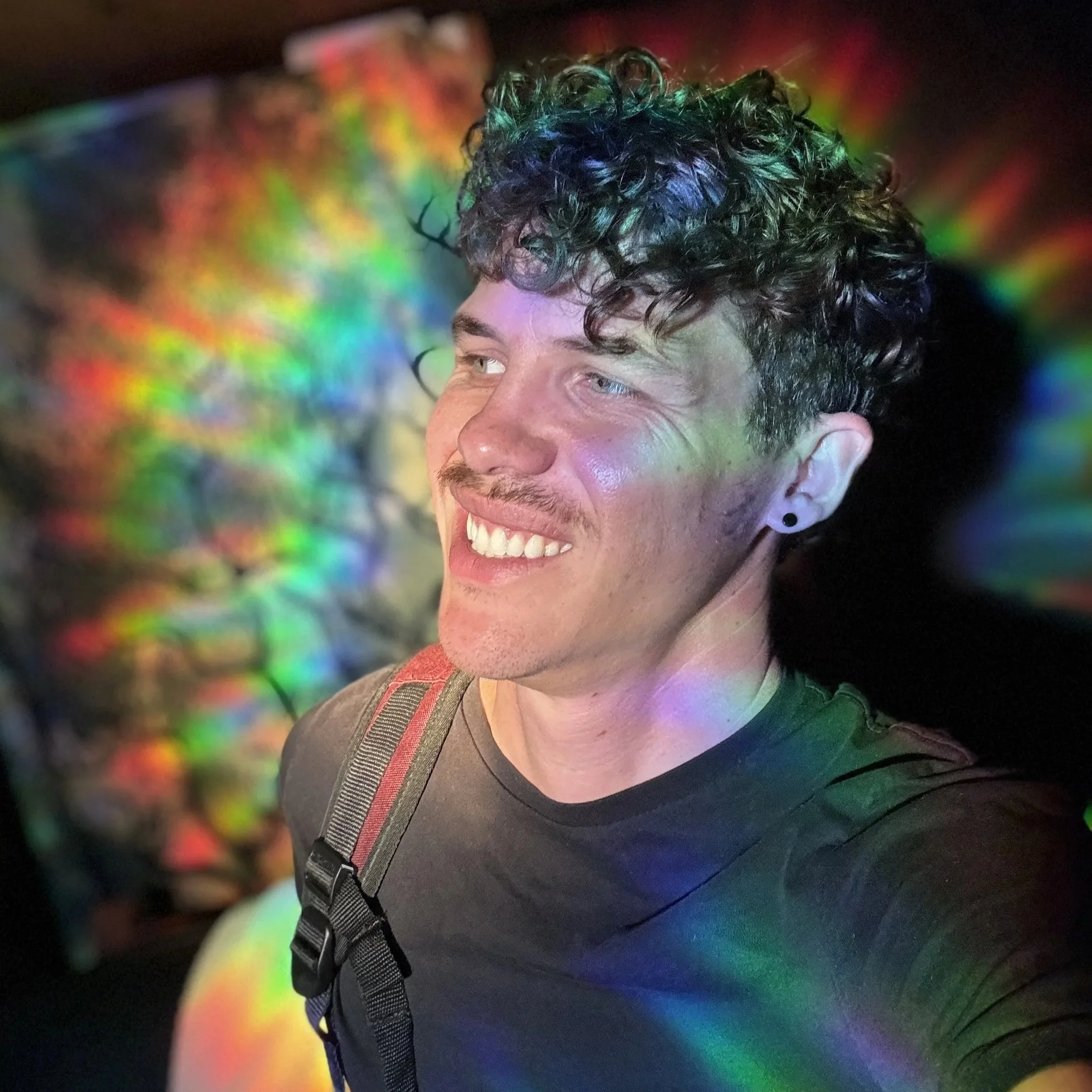 Sömmer is a gender queer white person with curly brown hair.  They have a black stud earing and t-shirt and are smiling and staring off to the viewer's left while awash in prismatic rainbow lighting.