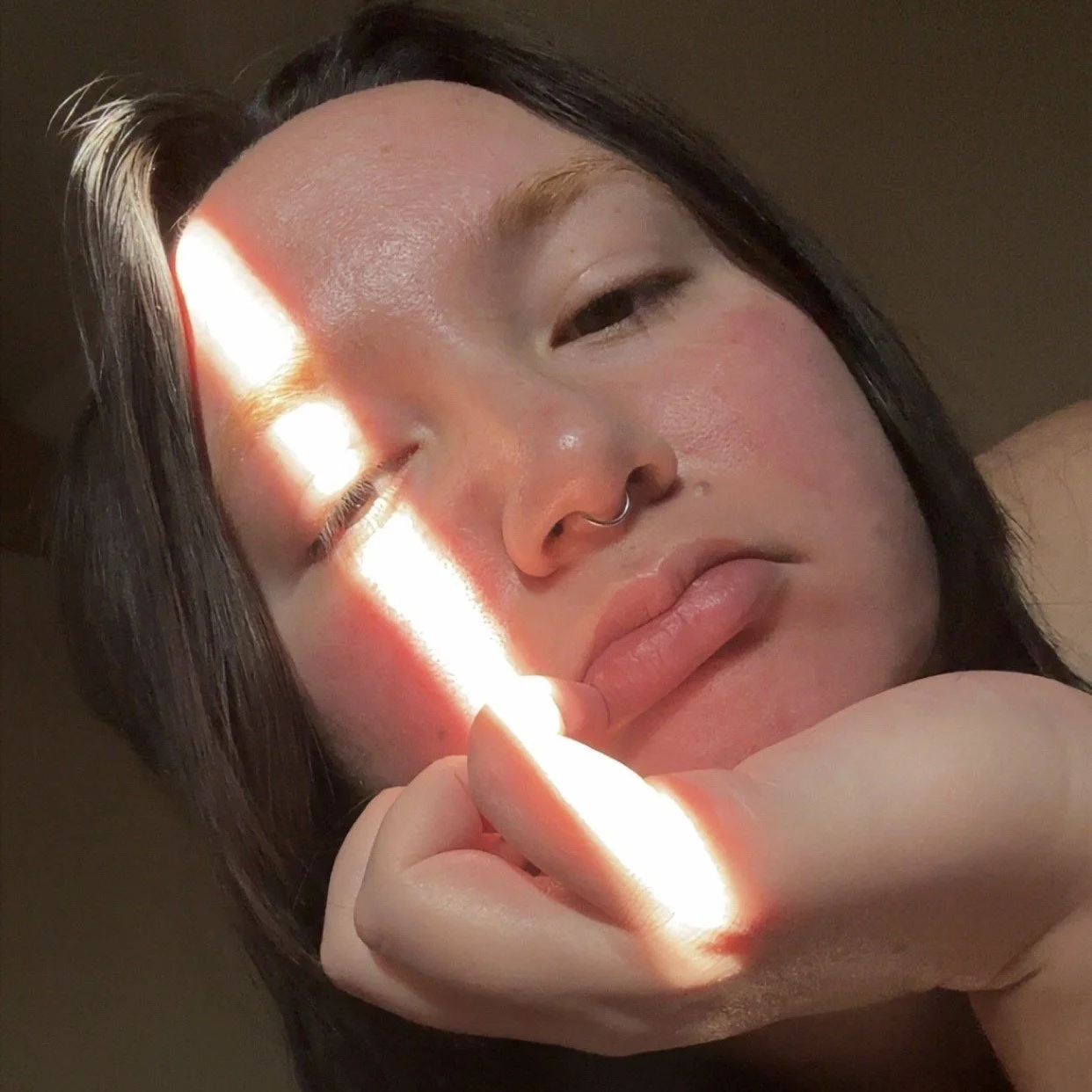 Hahana is a māhū ʻōiwi with dark brown shoulder length hair, bleached eyebrows, and a septum piercing. He is leaning his chin against his left hand and keeping a neutral facial expression. A sliver of light is catching their left eye.