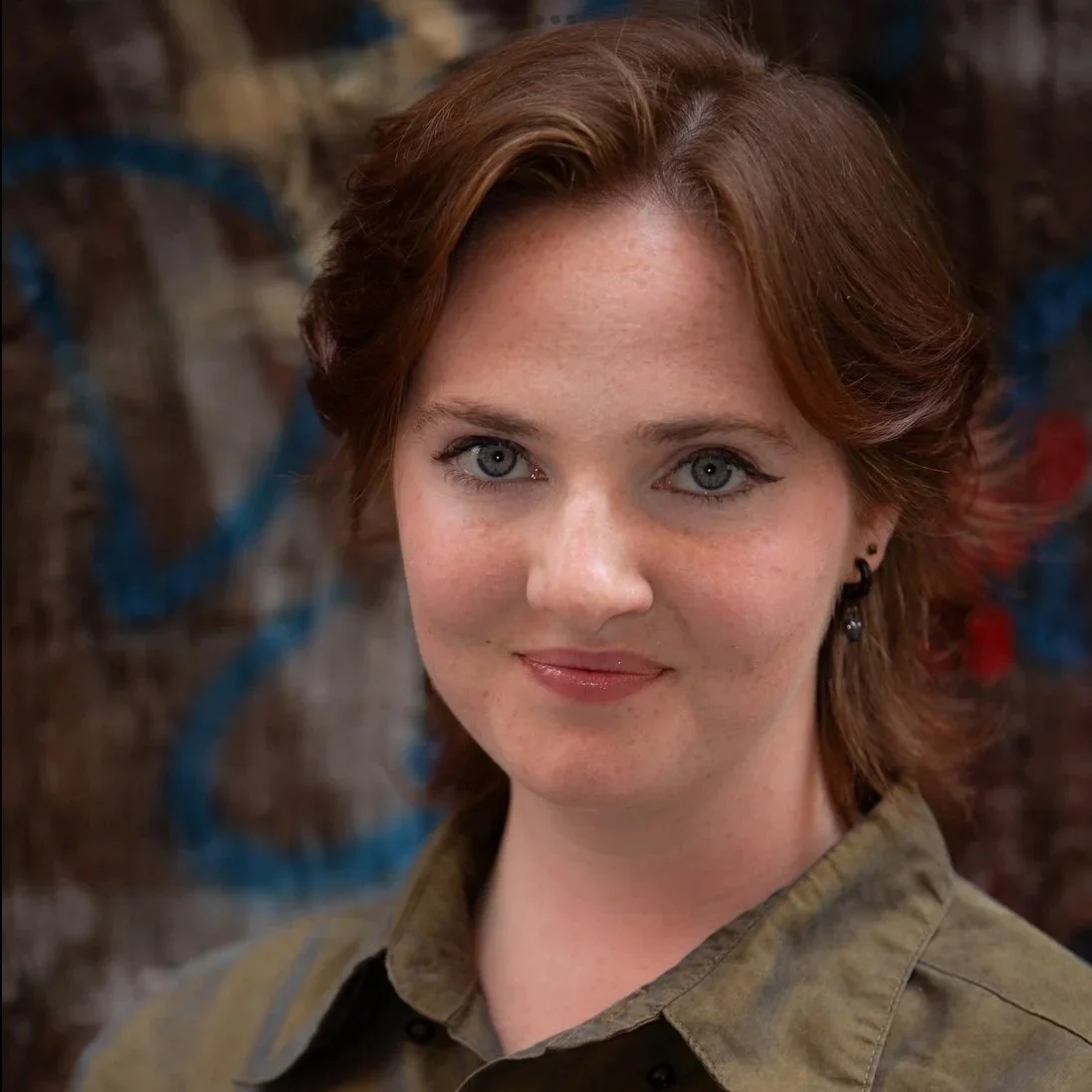 Karina is a white person with shoulder-length brown-red hair and blue-grey eyes. She is wearing a dark green button up shirt and smirking directly into the camera.Behind is street graffiti with blues and purples