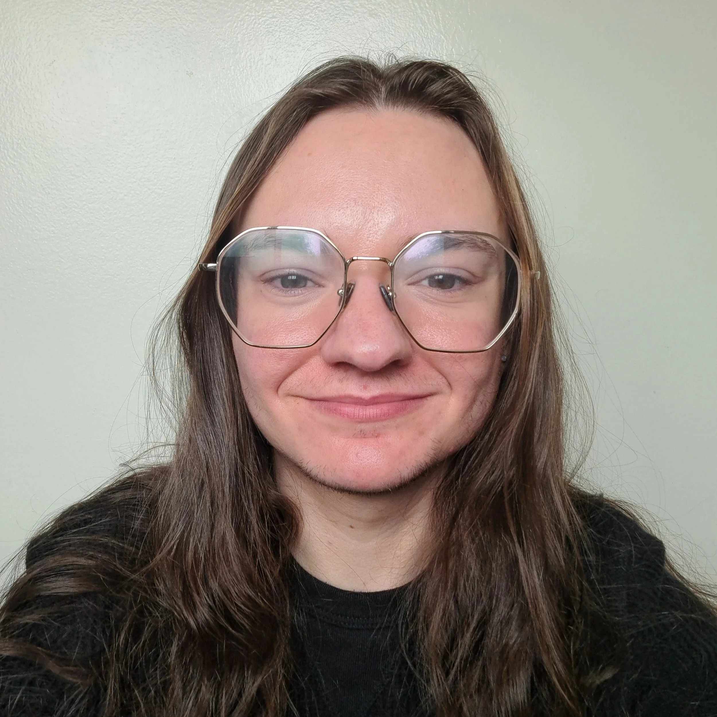 Mors is a transman with long brown hair. They are white, and are wearing a black sweater and silver glasses. Mors is smiling a small smile. The background is white.