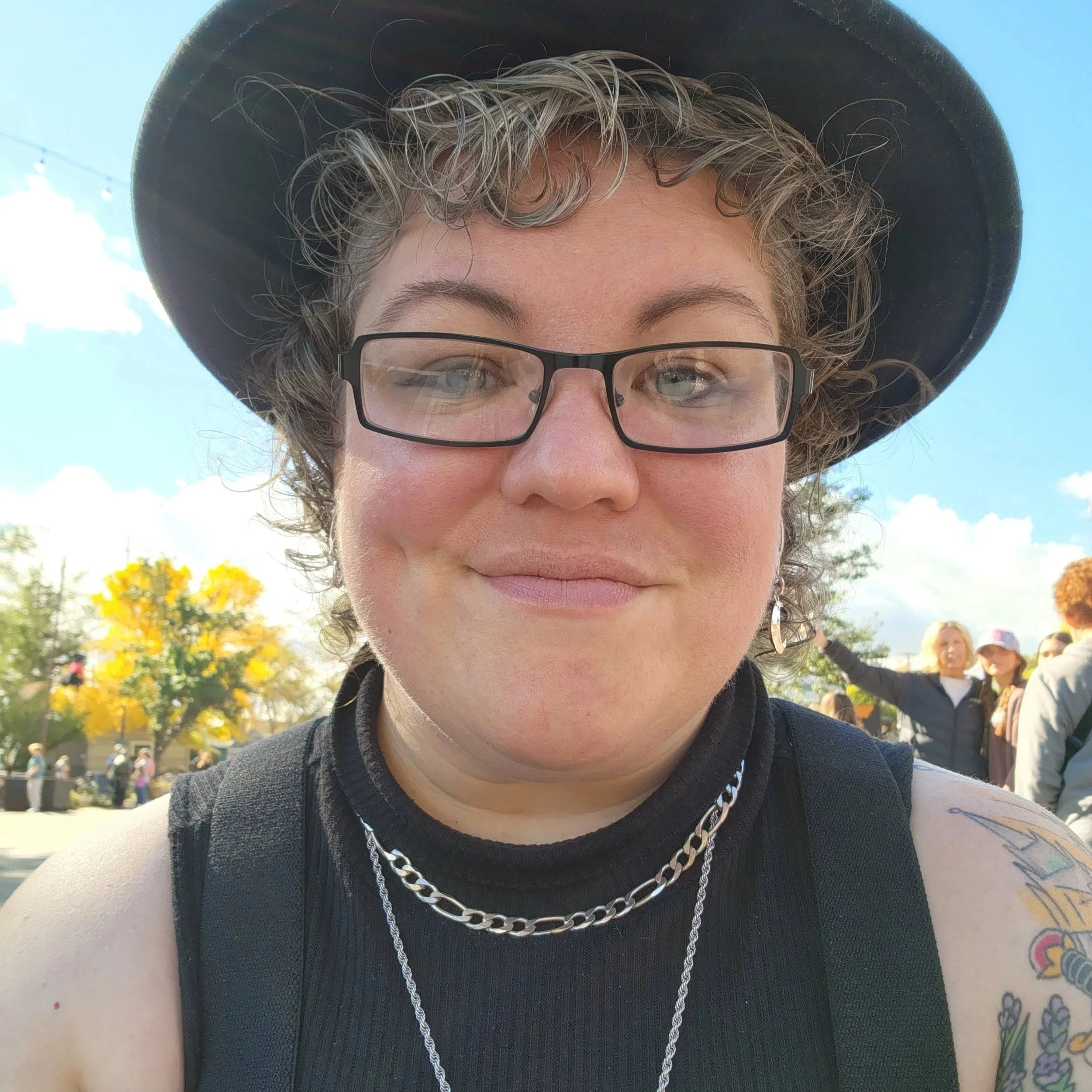 Kat is a non-binary person with curly brown and silver hair. They are wearing a wide-brimmed black hat, a black sleeveless turtleneck, and black suspenders and are smiling with a soft confidence. Behind is clear blue sky, autumn trees, a small crowd.