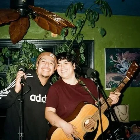 Two Latin trans men, smiling. Judas is wearing a caramel beanie, a black ADIDAS hoodie, holding a mic stand. Azul has dark curly hair, wearing a burgundy t-shirt, holding a guitar. Behind is a green wall with foliage, a brown ceiling fan.