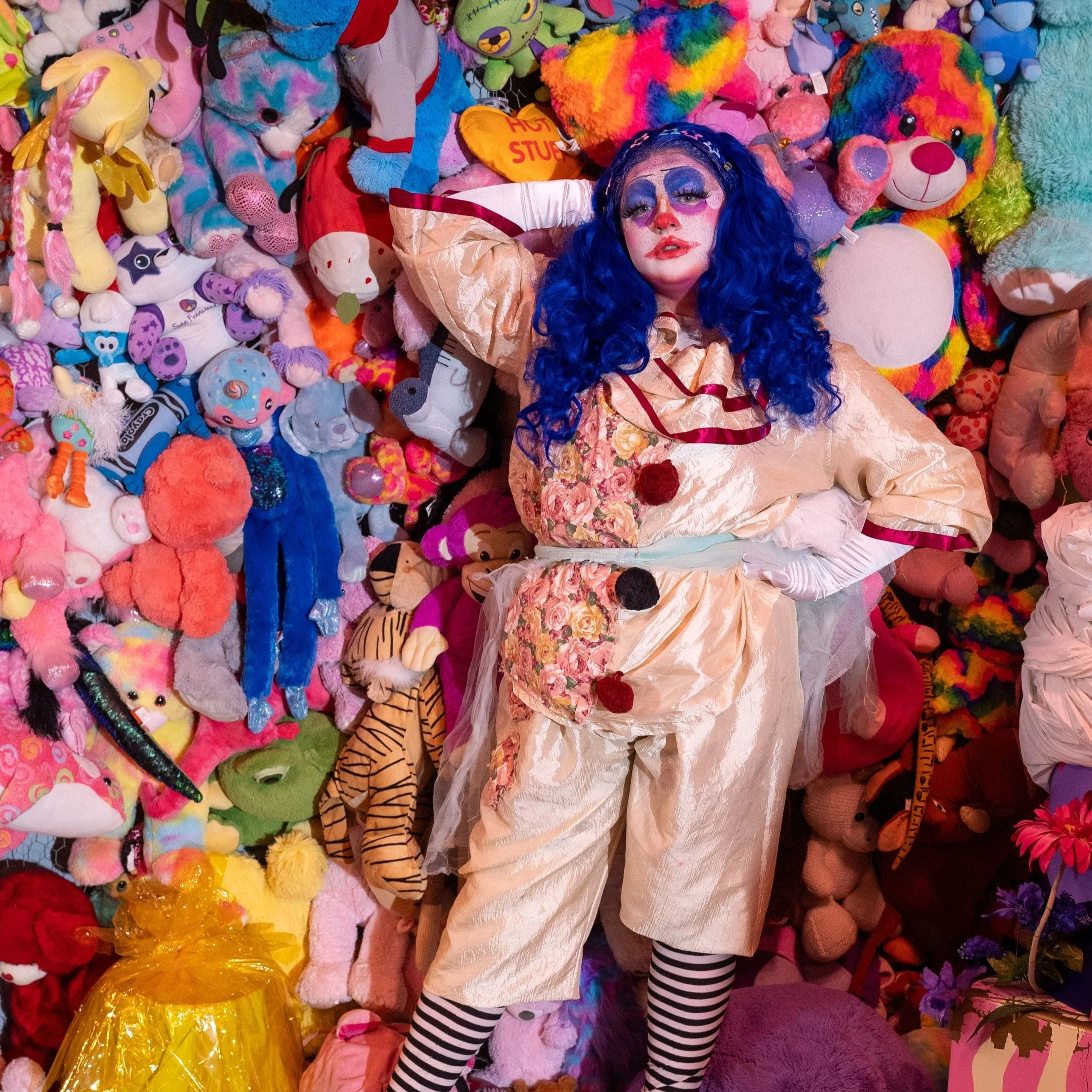 Shortcake the clown stands in front of a wall of plushies and stuffed animals, dressed in red and off-white colors. She wears bright pink platforms and has a curly long, blue wig that cascades down her shoulders contrasting with the white gloves.