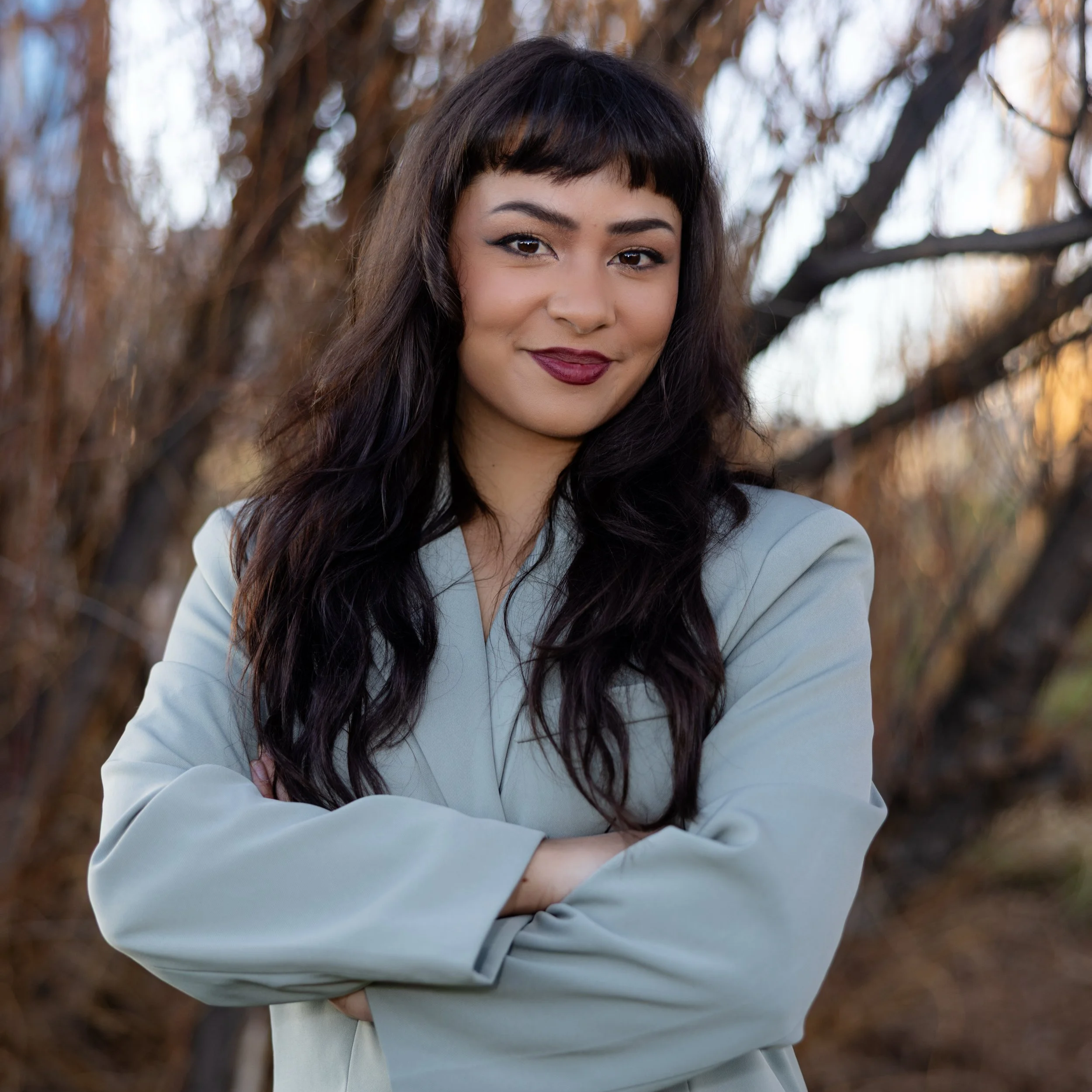 Denyse is a brown Filipina genderqueer woman in her early 30s with long brown hair and bangs. She is standing outdoors and smiling softly with her mouth closed. She is wearing a sage green suit and has her arms crossed and dark makeup.