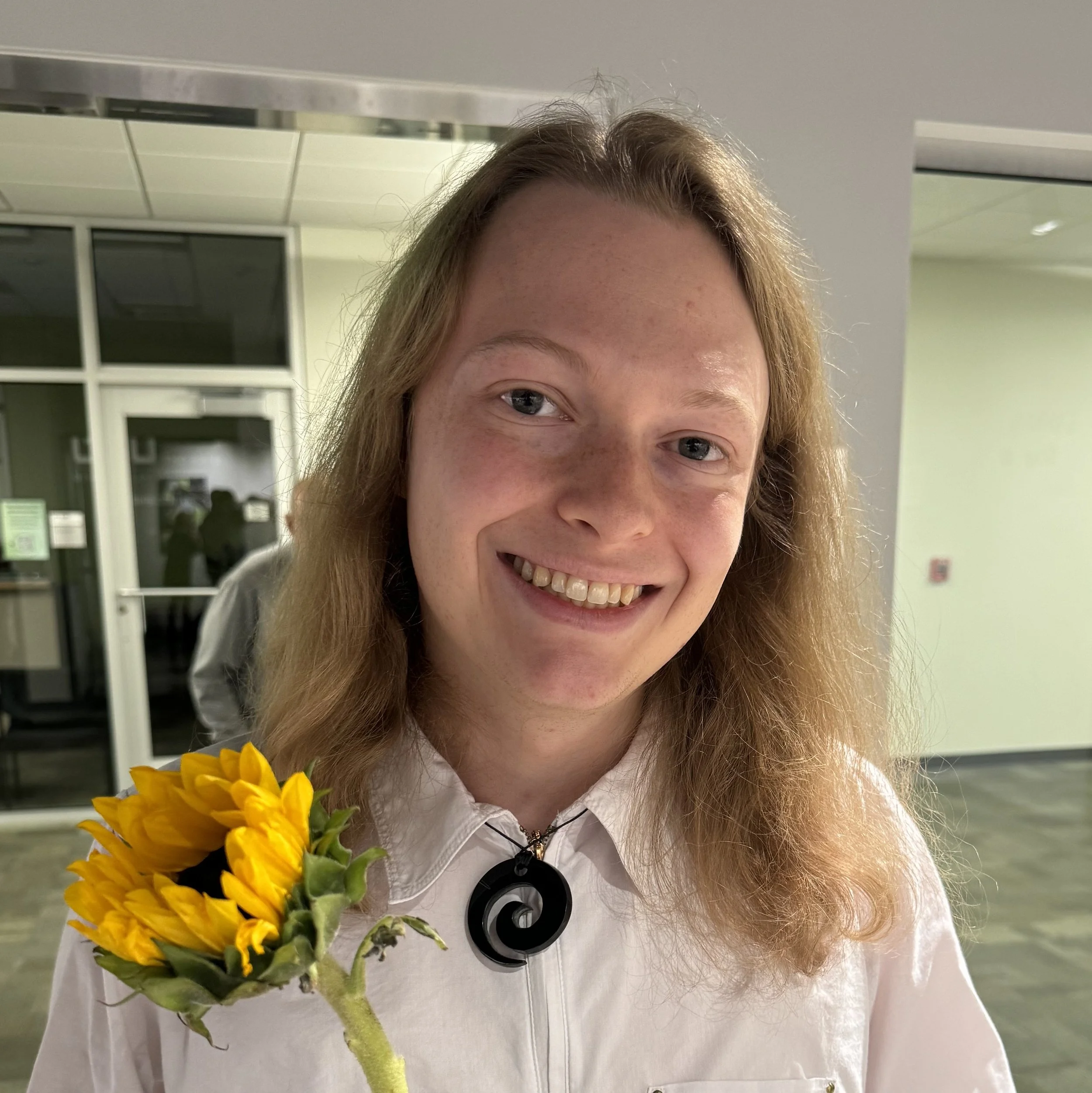 David is a queer white person with dirty blonde hair past their shoulders. They're wearing a white shirt and blue-green swirl-shaped necklace. They're holding a sunflower, tilting their head, and smiling. There's a hallway behind them.