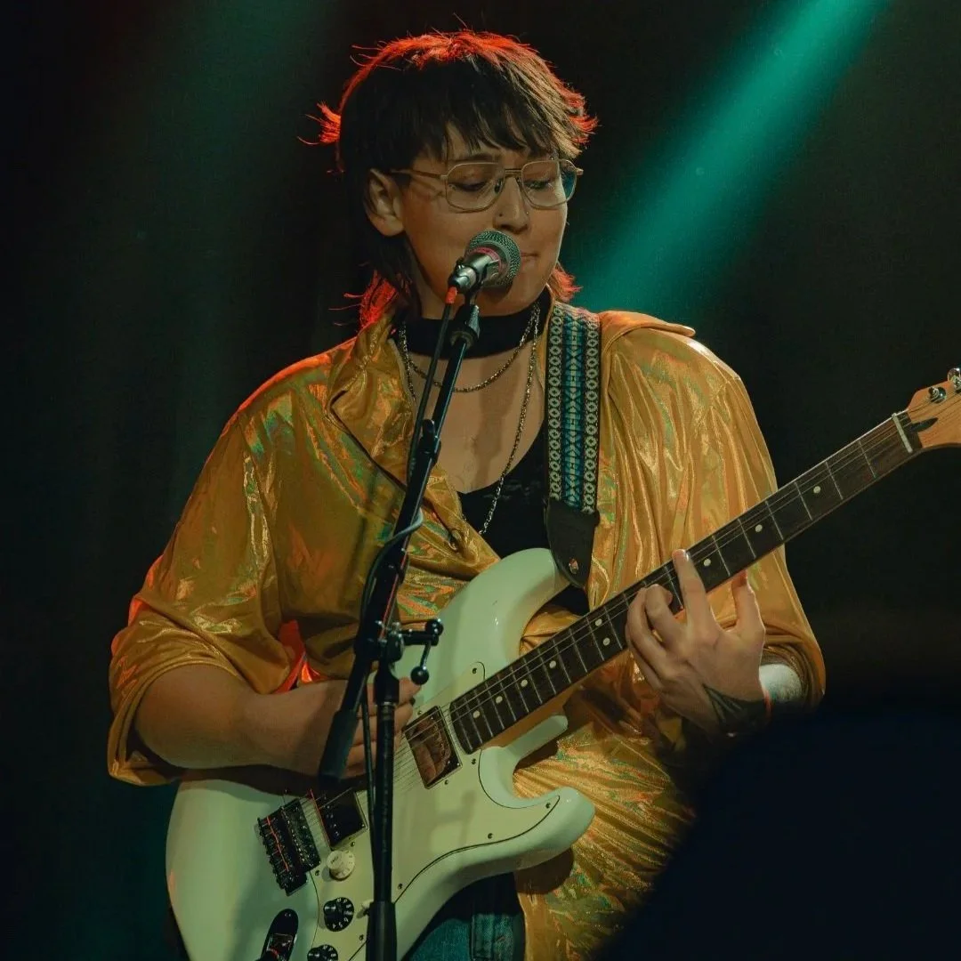 Moon is a gender queer white person with brown hair. Moon is on a stage, wearing a shimmery golden shirt, holding a guitar and singing into a microphone. Lights from the stage shine in the background.