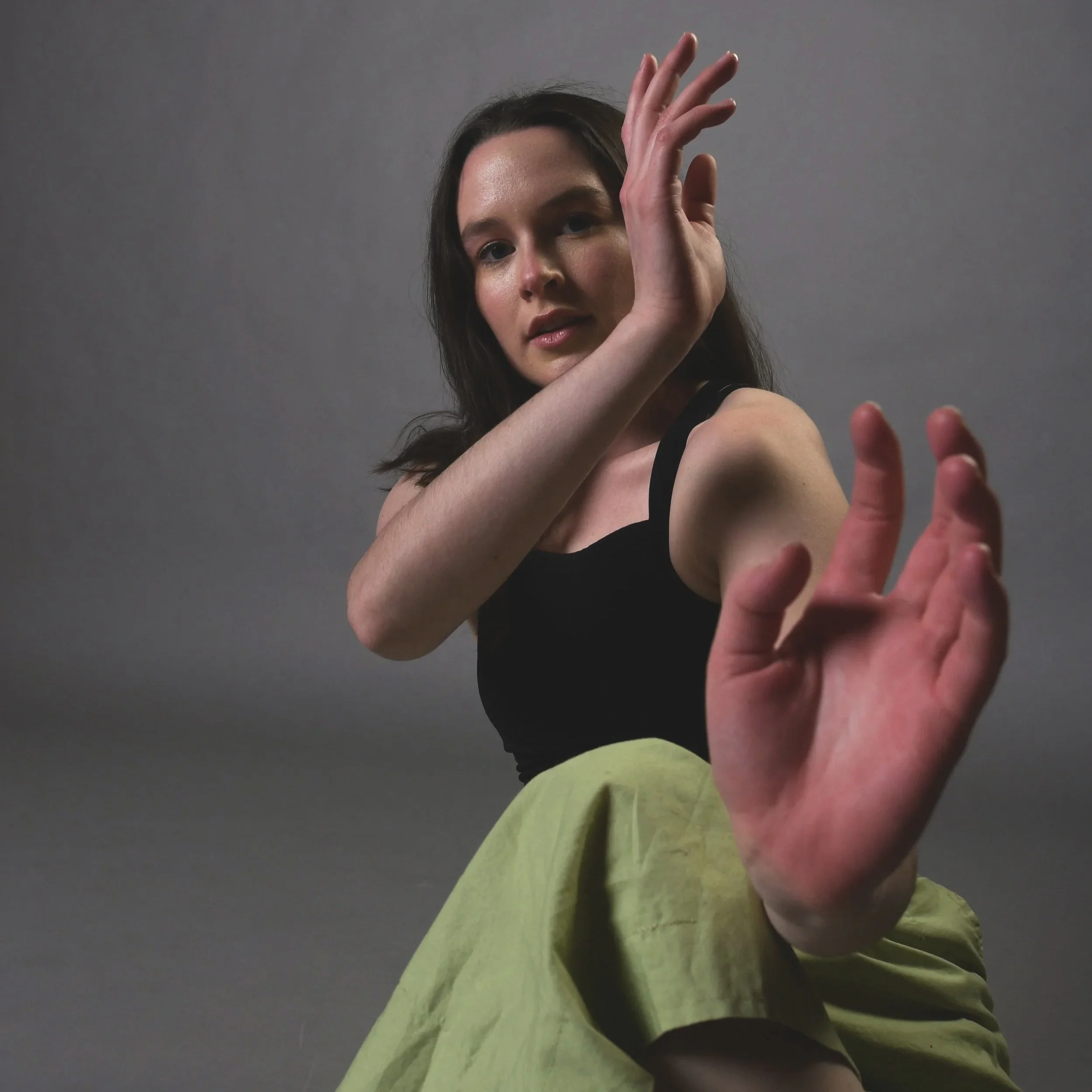 Kara is a feminine white person with long brown hair. She is wearing bright green pants and creating a shape with her arms that reaches to the camera.
