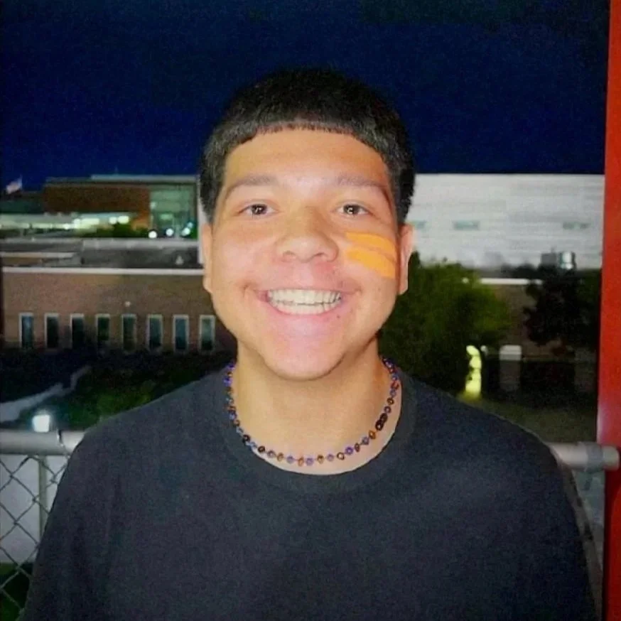 Isaiah is wearing a black shirt with a bead necklace and is smiling ear-to-ear. Isaiah has short black hair and is from Latin descent. A dark background set at night on top of bleachers is present.