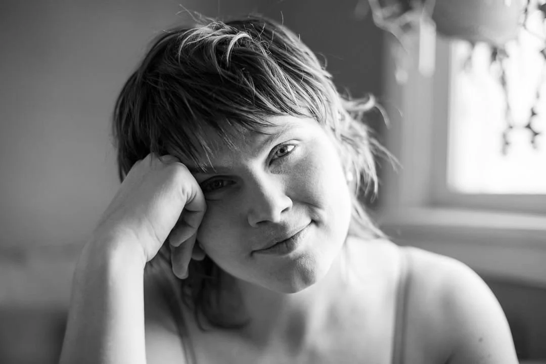 A closely-cropped b&w picture of Meagan, a white woman with soft shaggy hair, gazing into the camera and smiling softly, resting her temple against her closed hand. Light pours in across her face from the window behind her, in which hangs a plant.