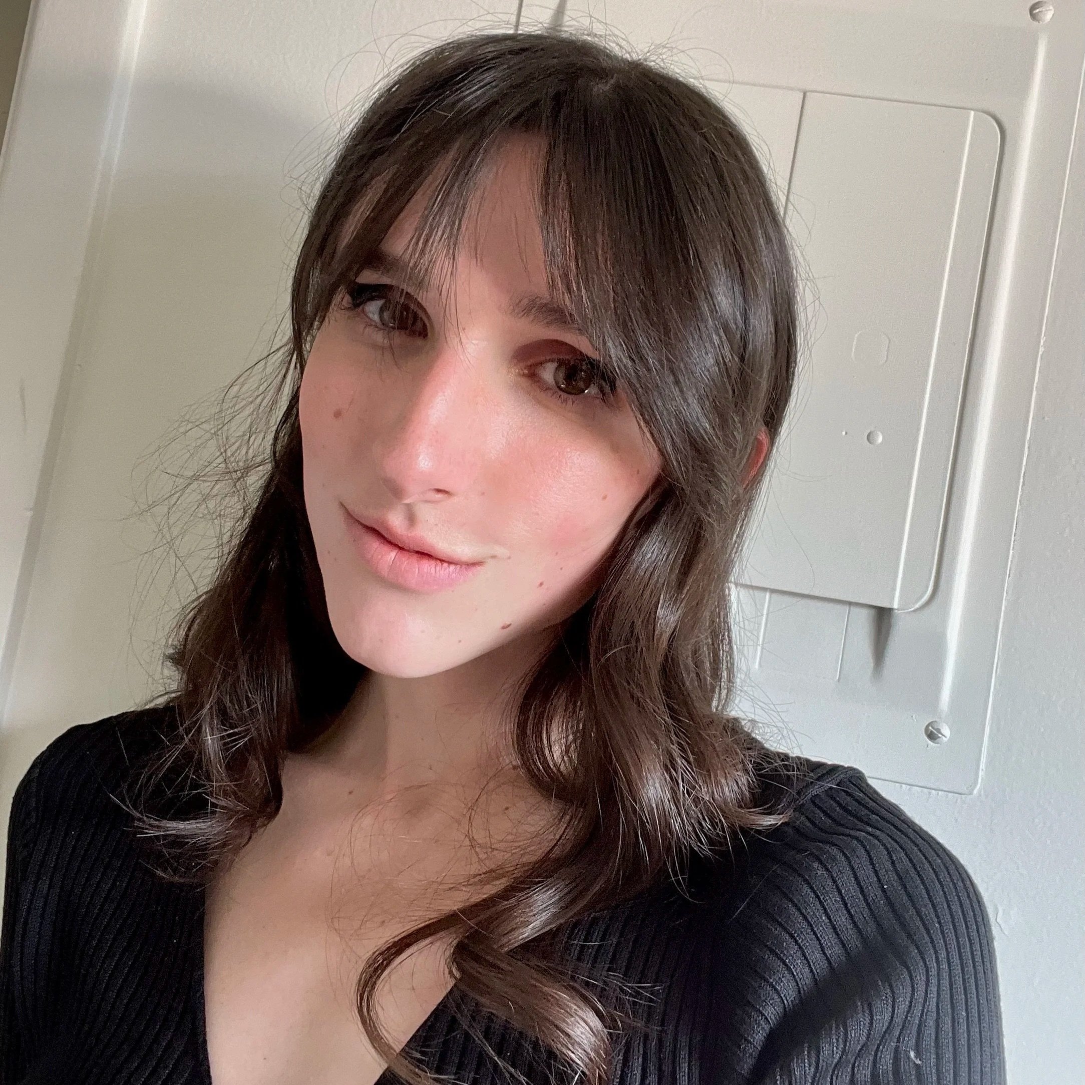 Joyce is a white woman with long dark hair set with subtle curls at the bottom. She is smiling and wearing a black long sleeved crop top in front of a blank wall.