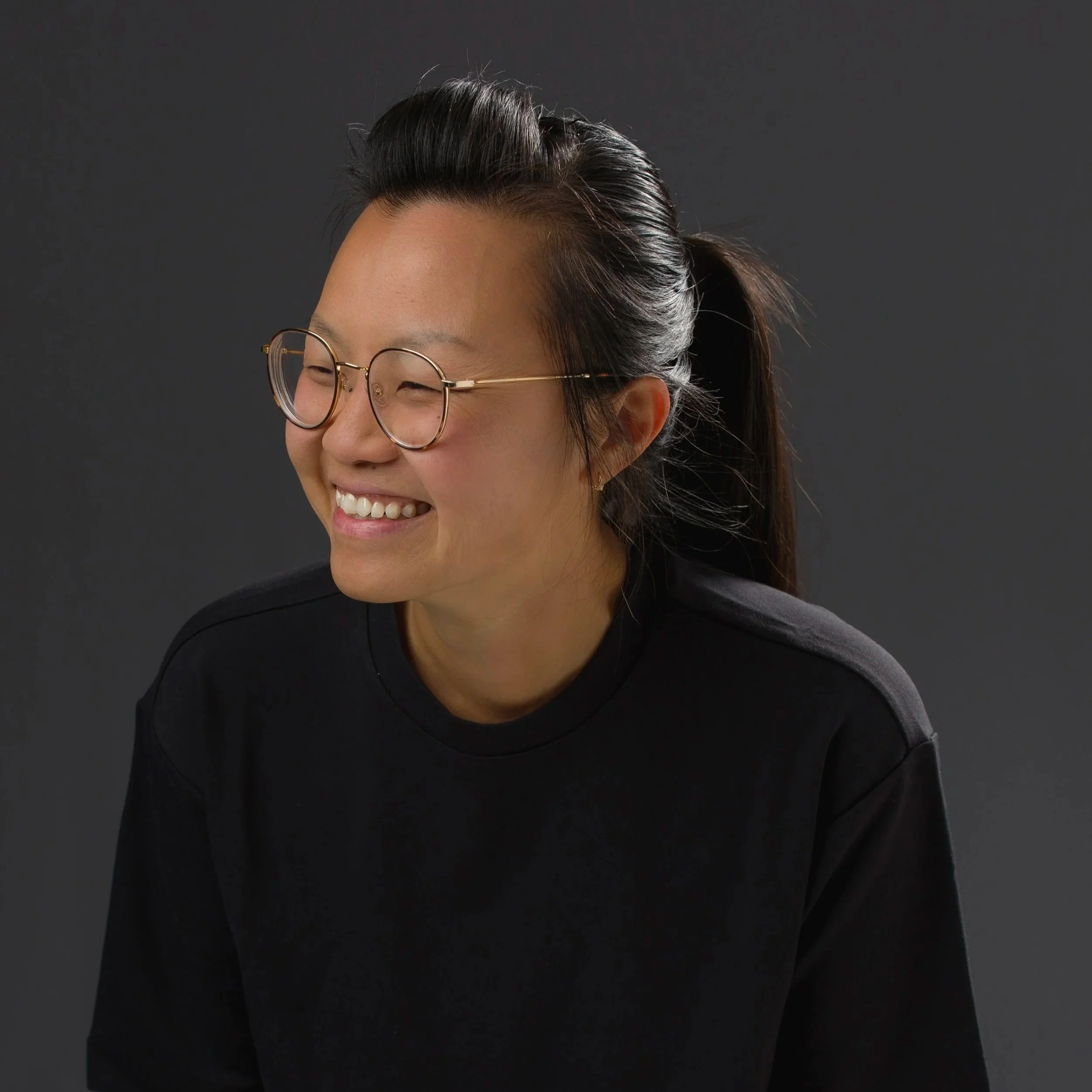 Thi is a gender queer person. in this picture, she has has a ponytail and is wearing a black top, browline style glasses, with a big smile. T stands against a dark grey background.