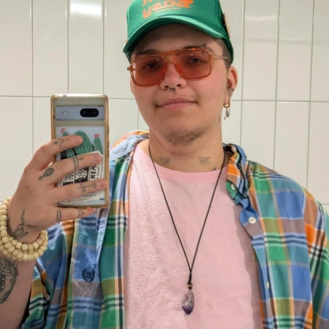 Beaux is a gender queer white person with a green trucker cap that says "Take it Easy", an amethyst necklace, and bone mala bead bracelet. They're wearing orange aviators, a pink t-shirt, and an oversized blue orange and green plaid button-up.