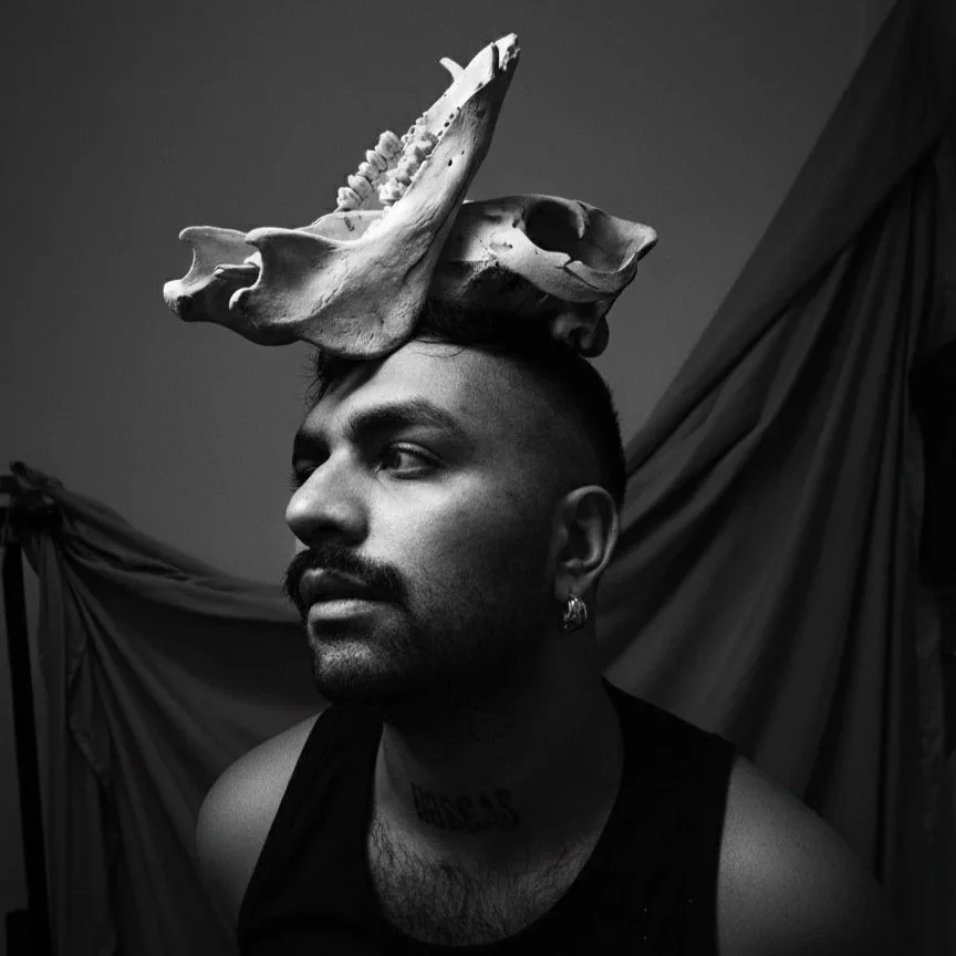 A black and white photo of a person with short dark hair and trimmed beard, turned slightly to the side. They are wearing a black sleeveless top and hoop earing with a large animal jaw bone positioned like a crown on top of their head. qqqqqqqqqqqqqq