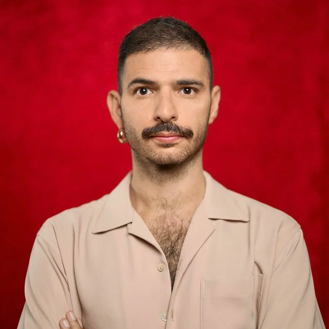 Hadi is a queer arab person with short hair, a little Mustache like freddie mercury, wearing a baby pink shirt and a little gold earring on his right side. Behind him is a red velvet background.