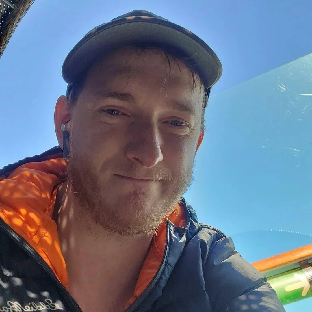 Hunter is sitting in a shaded area wearing a black and orange windbreaker and smiling softly in front of a sunny blue sky. He's wearing a baseball cap and has dirty blonde hair and a short beard.