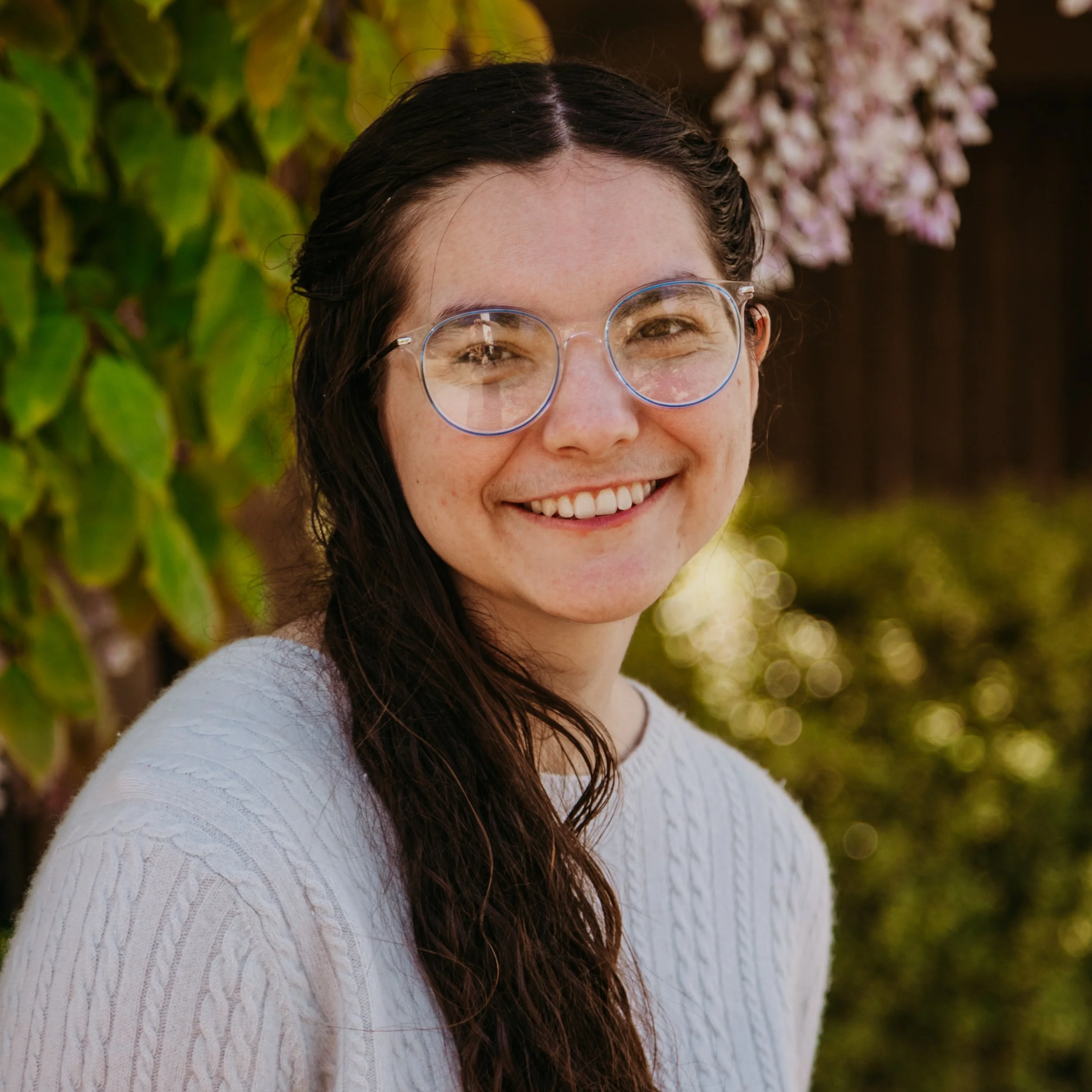 Ellie is a genderfluid white person with long dark brown hair. They are wearing a light blue sweater, round glasses with a clear rim, and they are smiling. There are leaves and flowers in the background.