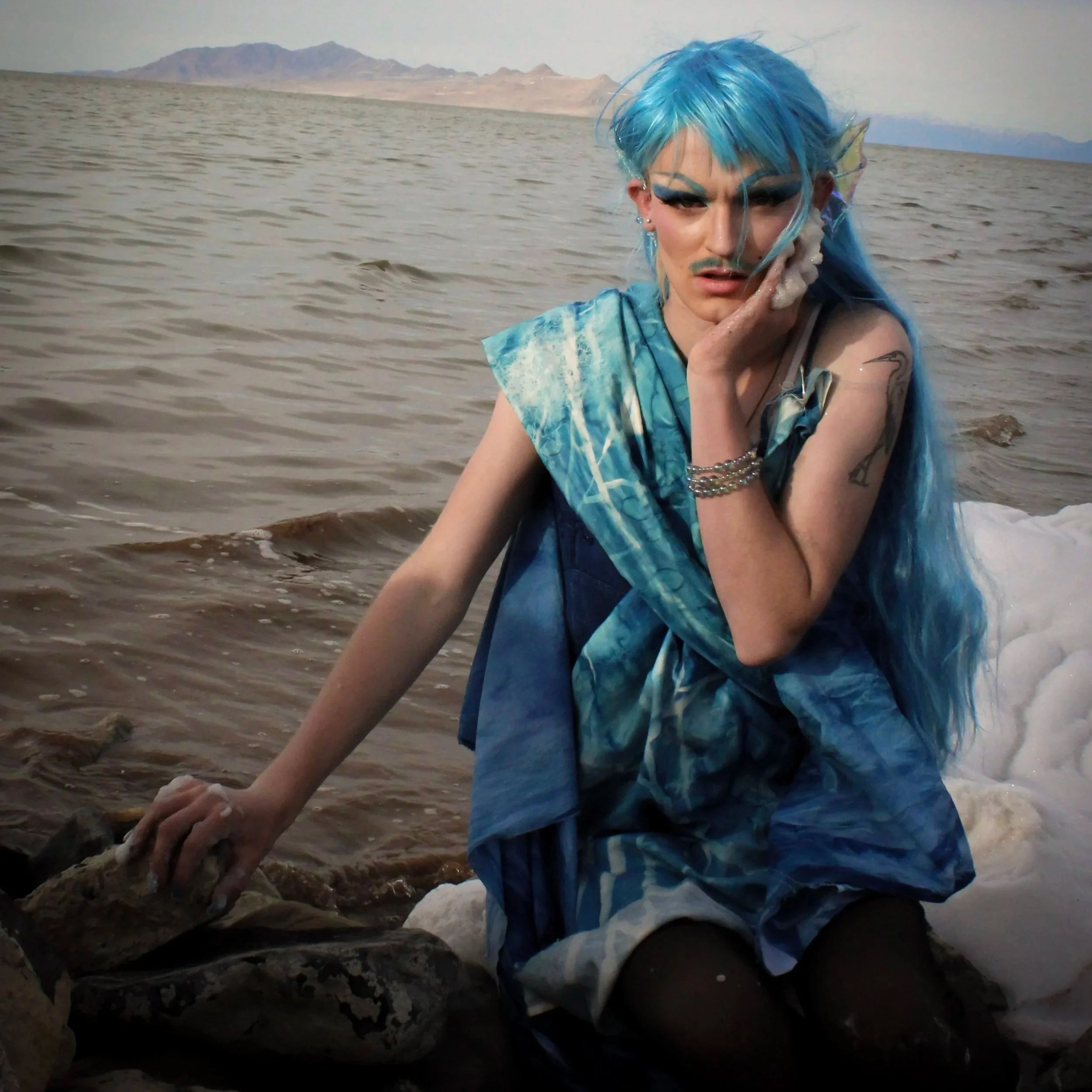Salina Marina is a white drag queen with blue hair. She has strong contour, blush, blue eyebrows, and dramatic eye makeup. She is draped sun-printed blue fabric and black tights. She is sitting on rocks on the shoreline of Great Salt Lake.