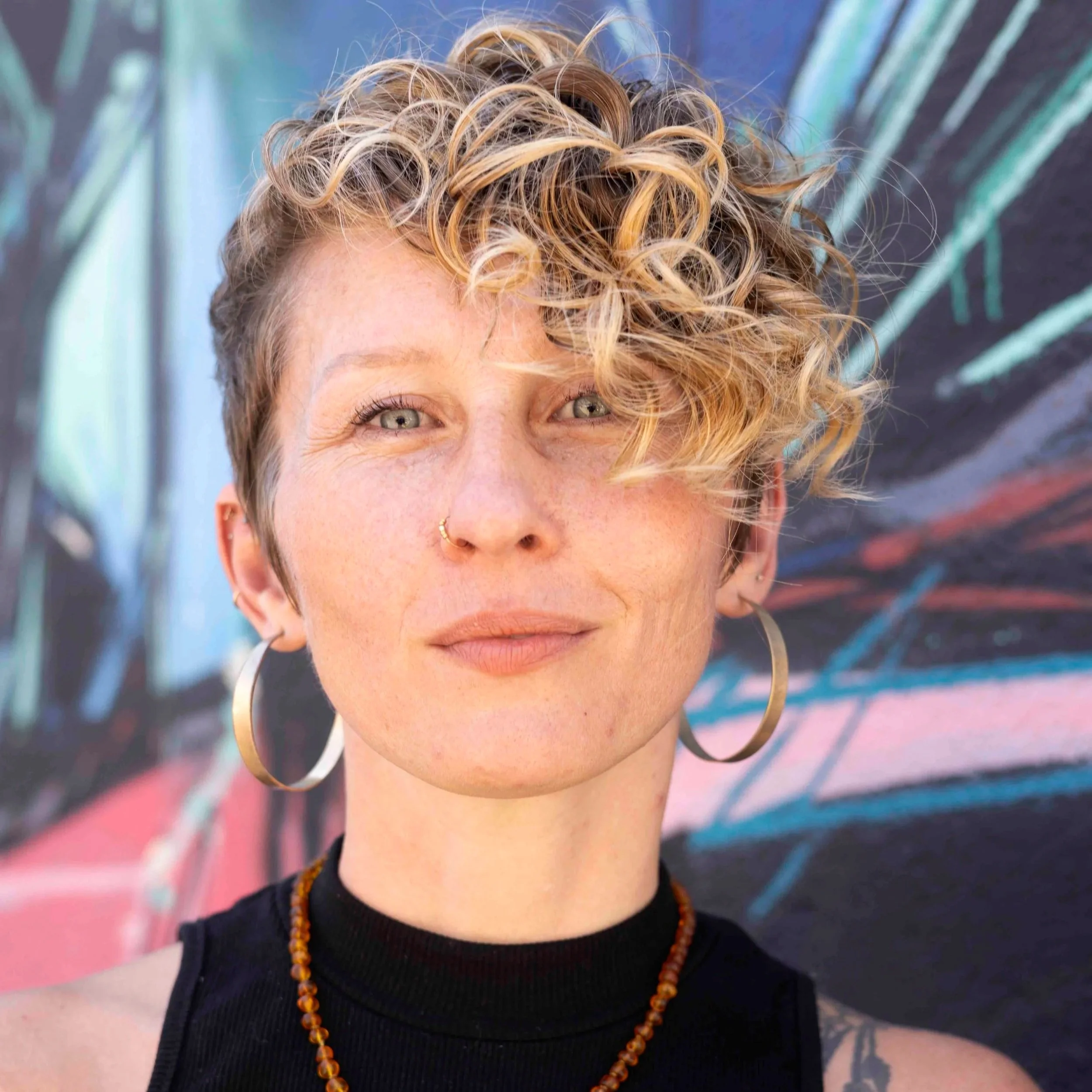 River is a genderfluid white person with short, curly, blond hair. River has brilliant blue eyes and a coy smile. They are wearing large hoop earrings, a small amber necklace, and a black sleeveless mock-neck shirt.