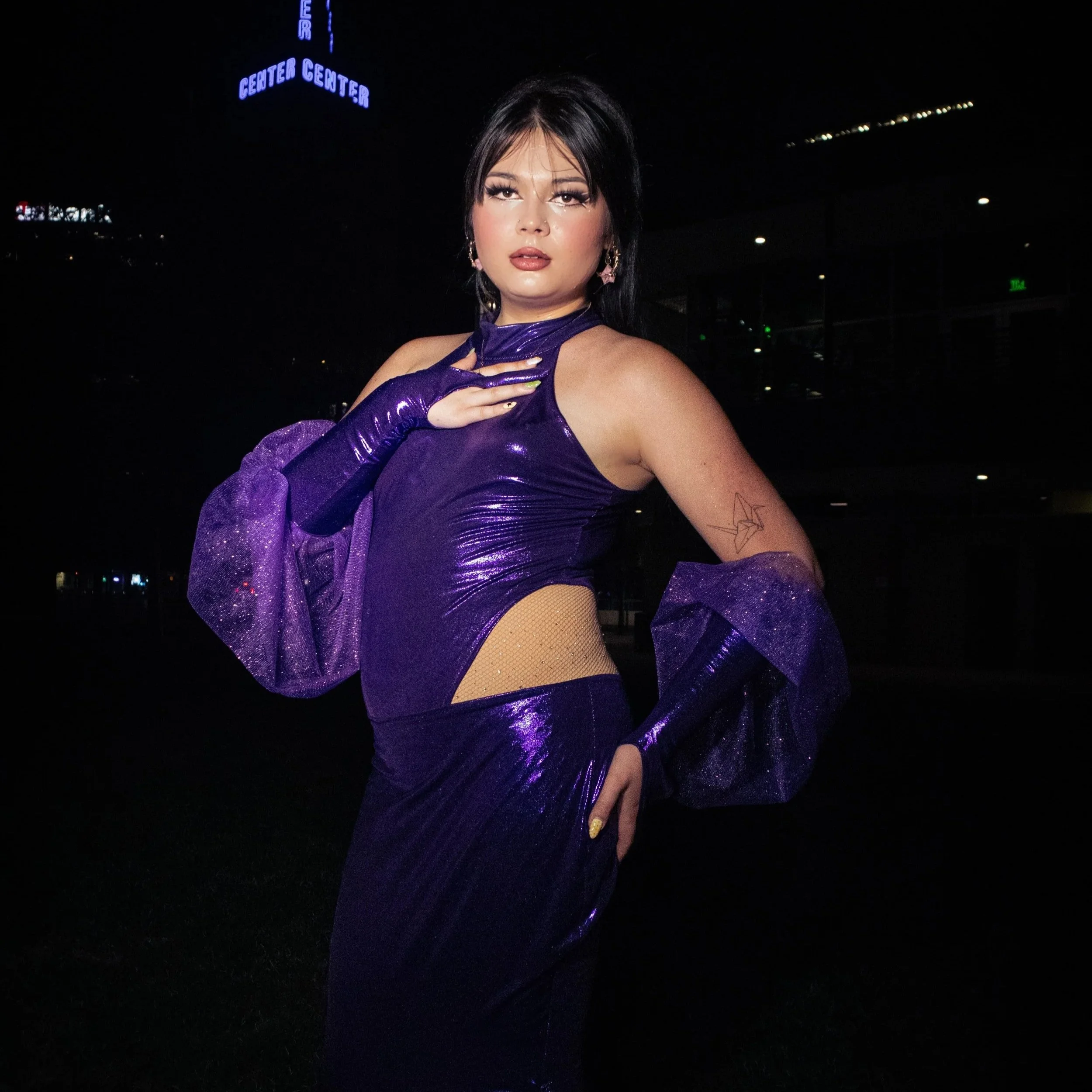 Aiko is a Japanese-American Trans woman with a long brown ponytail. Aiko is wearing a purple, sparkly mermaid-tail gown with matching gloves. She is looking into the camera with intention, with a dark, city background behind her.