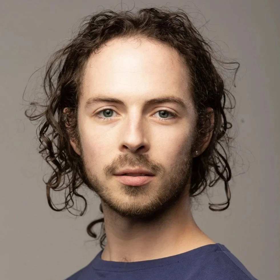 Tyler is a genderqueer white person with mid-length curly brown hair. They have facial hair and are wearing a blue shirt with a grey background.
