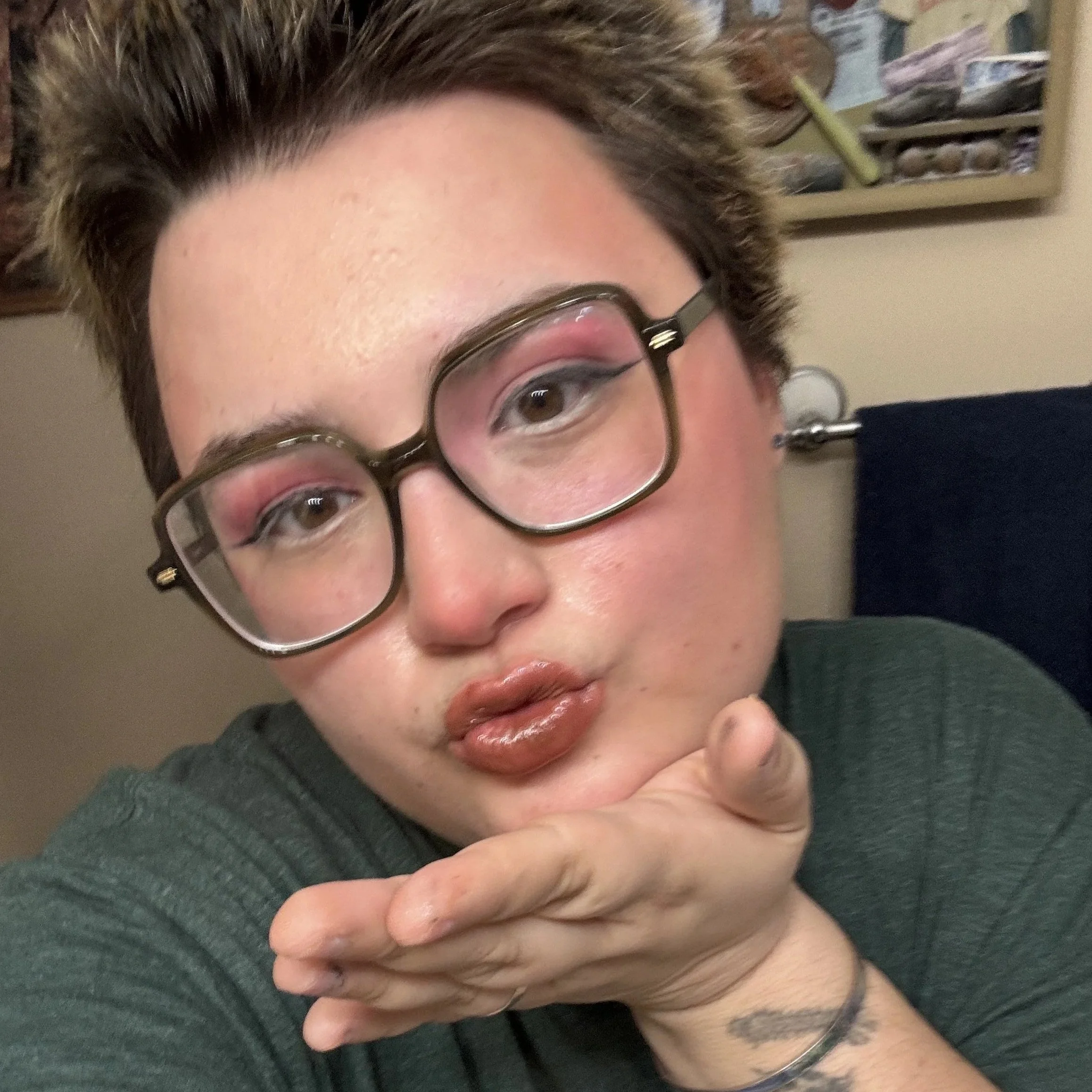 Emeline is a queer white person with shaved brown hair wearing large-frame, black glasses.  She is blowing you a kiss! You'd better catch it.