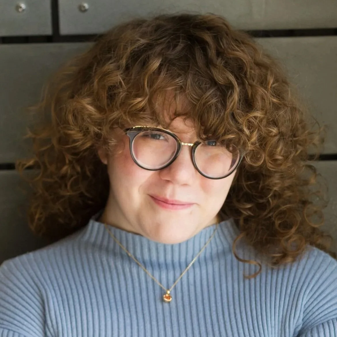 Morgan is a white nonbinary person with shoulder-length curly brown hair, wearing black and gold glasses, a blue ribbed sweater, a gold pomegranate necklace with red jewels. They are smiling with their mouth closed. Background: dark gray-green planks