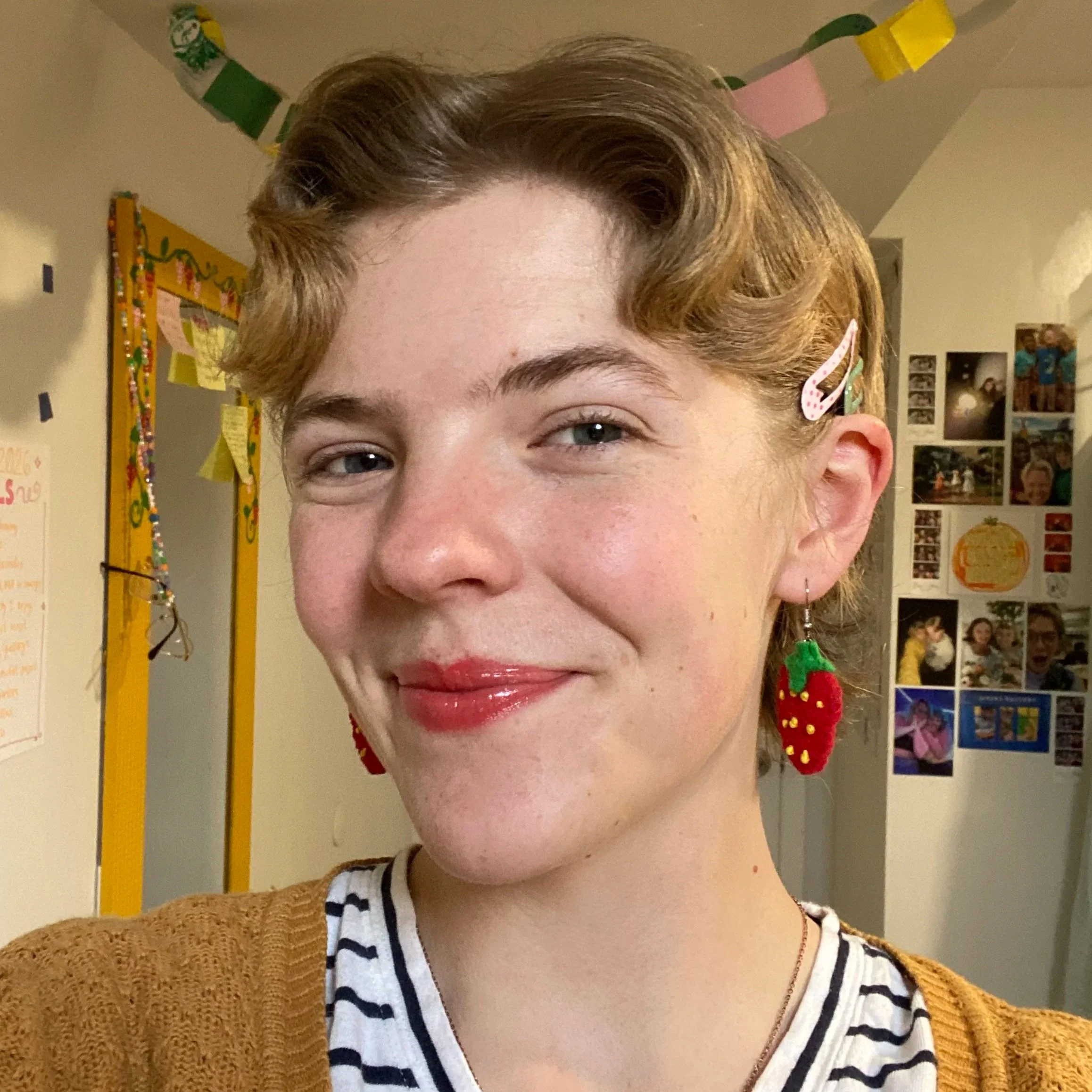Hannah is a white woman with short blonde hair. She is wearing strawberry earrings, a black and white striped shirt and a yellow cardigan