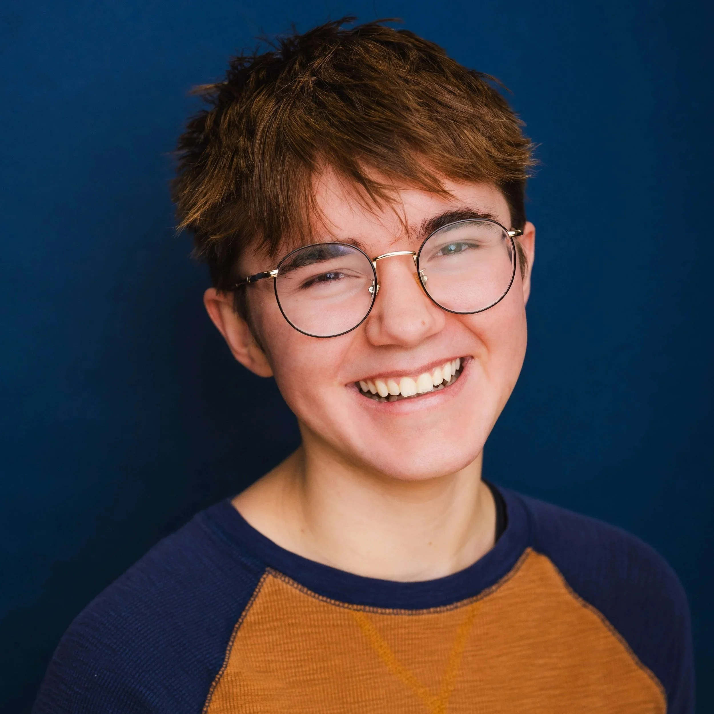 Evan is a white transgender man with short brown hair. He is wearing round gold glasses and in a ochre yellow shirt with navy blue sleeves. He is against a blue background. He's smiling widely with his head tilted slightly.