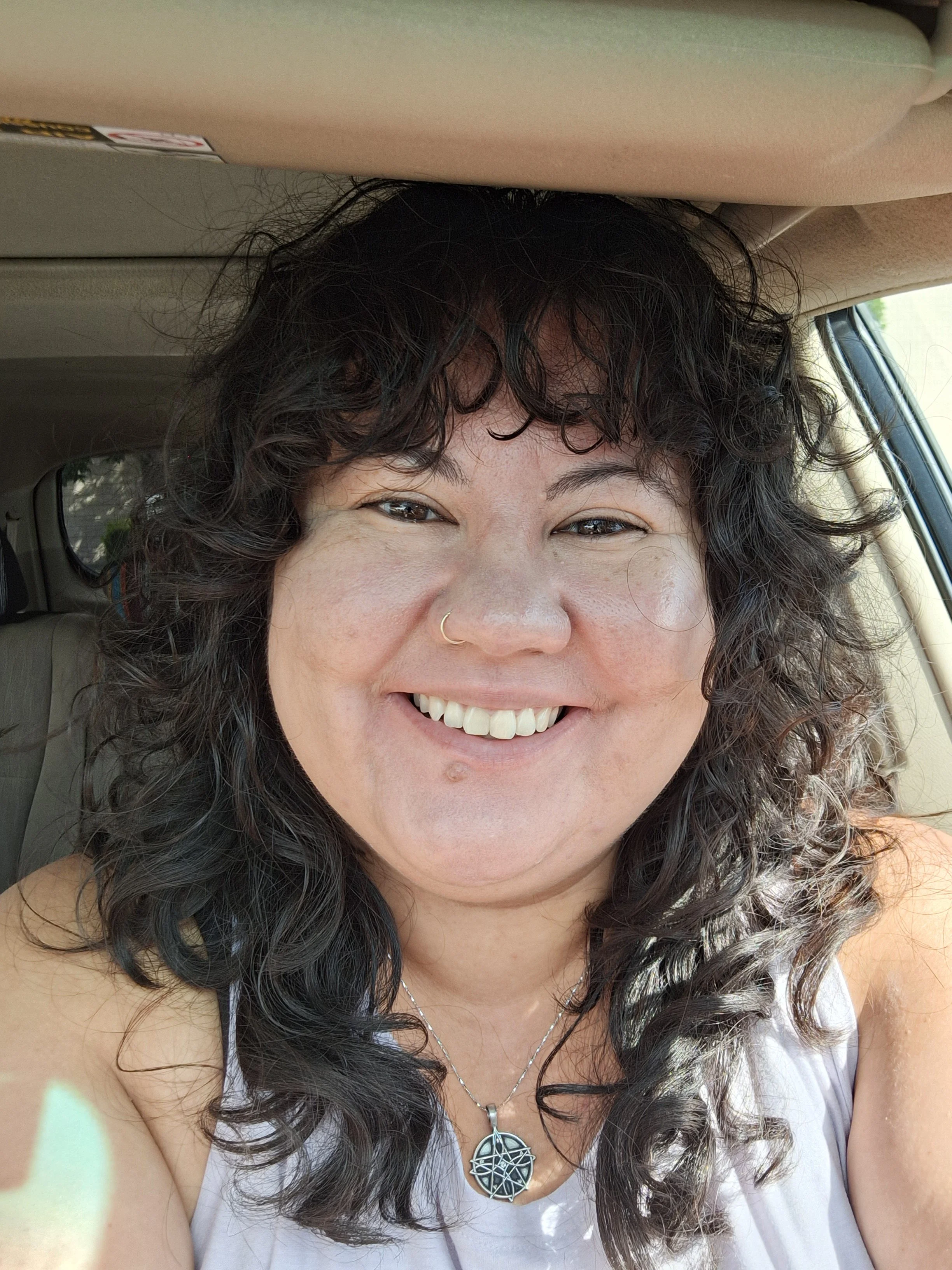 Sarah is a queer brown woman with curly brown hair. Sarah is wearing a blue demin jacket with a multi-colored scarf. She is sitting in her car with the carseats visible in the background.