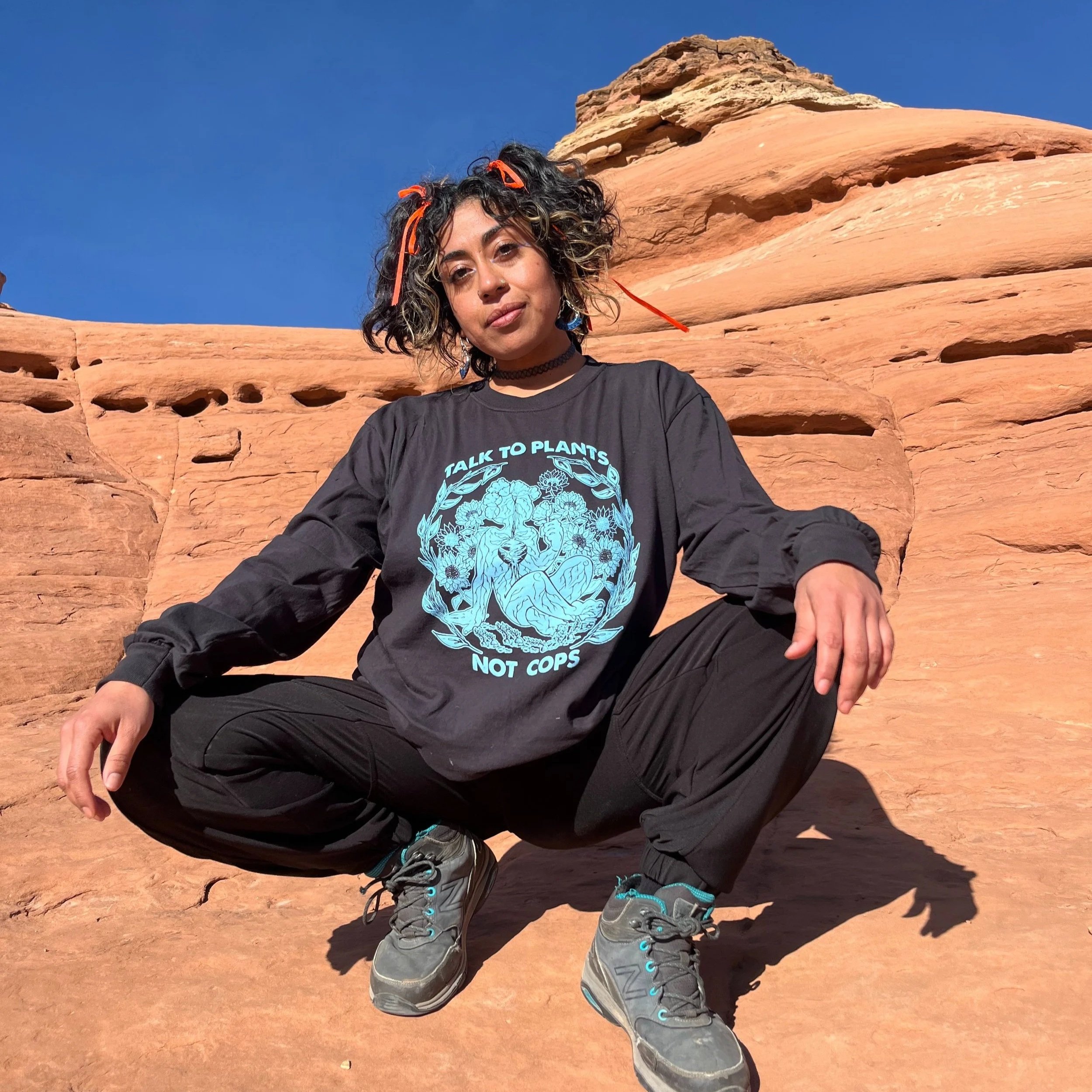 cesia is a cuir Mixteca with cinnamon skin and short curly black hair with neon orange ribbons in their hair. They are wearing a shirt they silk-screen that says “talk to plants not cops” as they squat on Arches red canyon rocks.