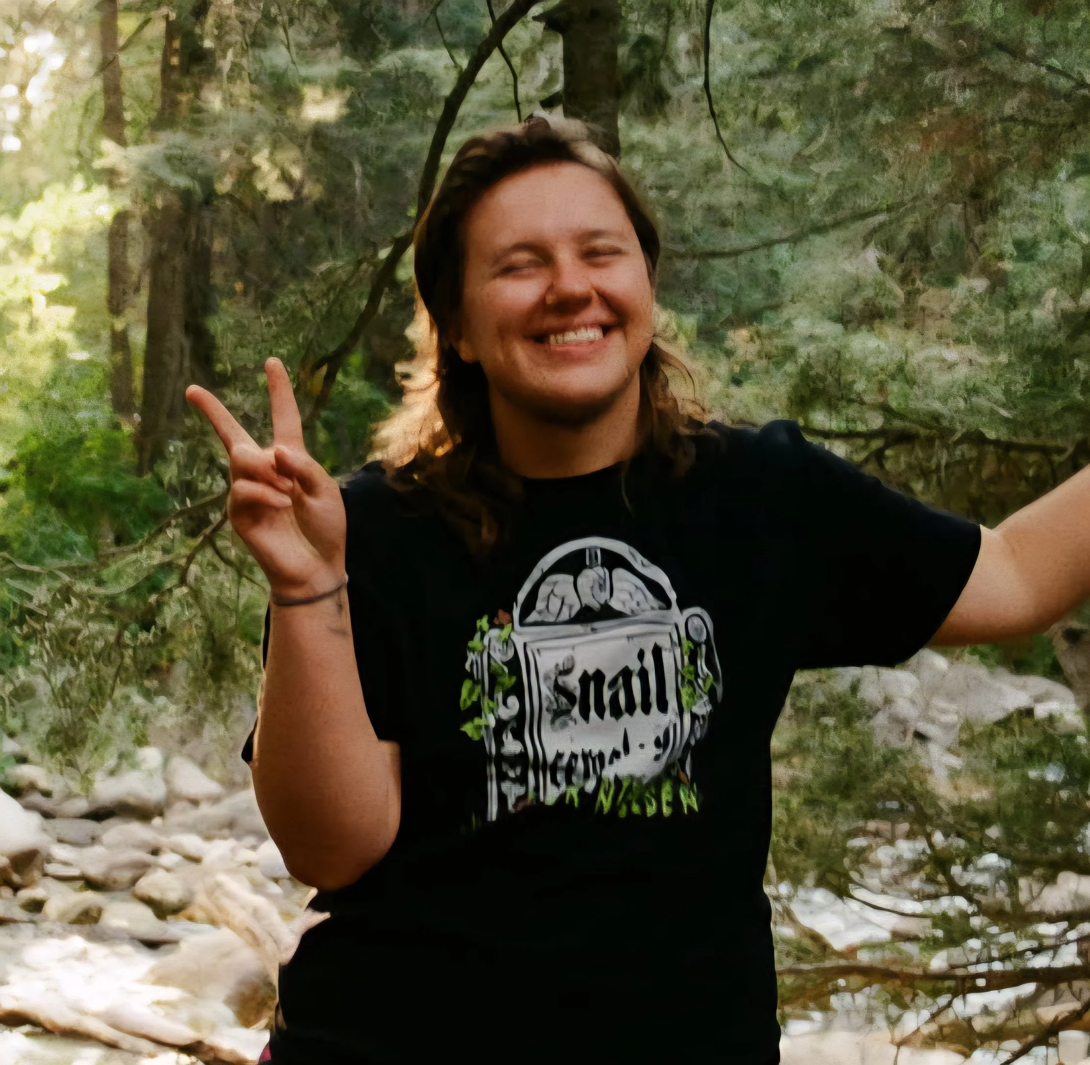 Kade is a non-binary, white trans man with shoulder length brown hair. He is smiling and holding up a peace sign. He is standing in the woods and wearing a black shirt with a gravestone on it that says 'Snail Cemetery'.