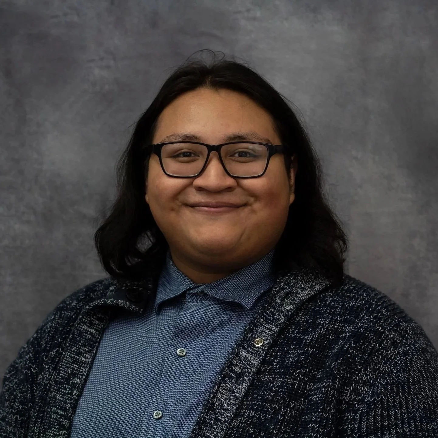 Pablo is a cis gender queer male person with glasses and long dark hair. Wearing a knitted overcoat sweater over a light blue button up, in front of a neutral grey blue background. Wearing dark rimmed square glasses frames.