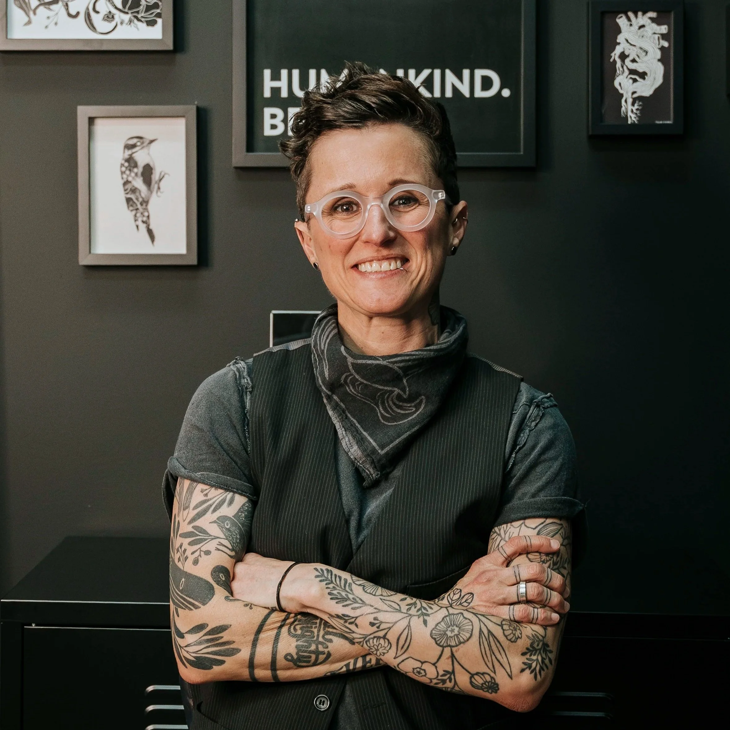 Nicole is a queer white person with short brain hear. She's wearing all black, a bandana and black glasses. She's in her tattooing studio with artwork on the wall behind her.