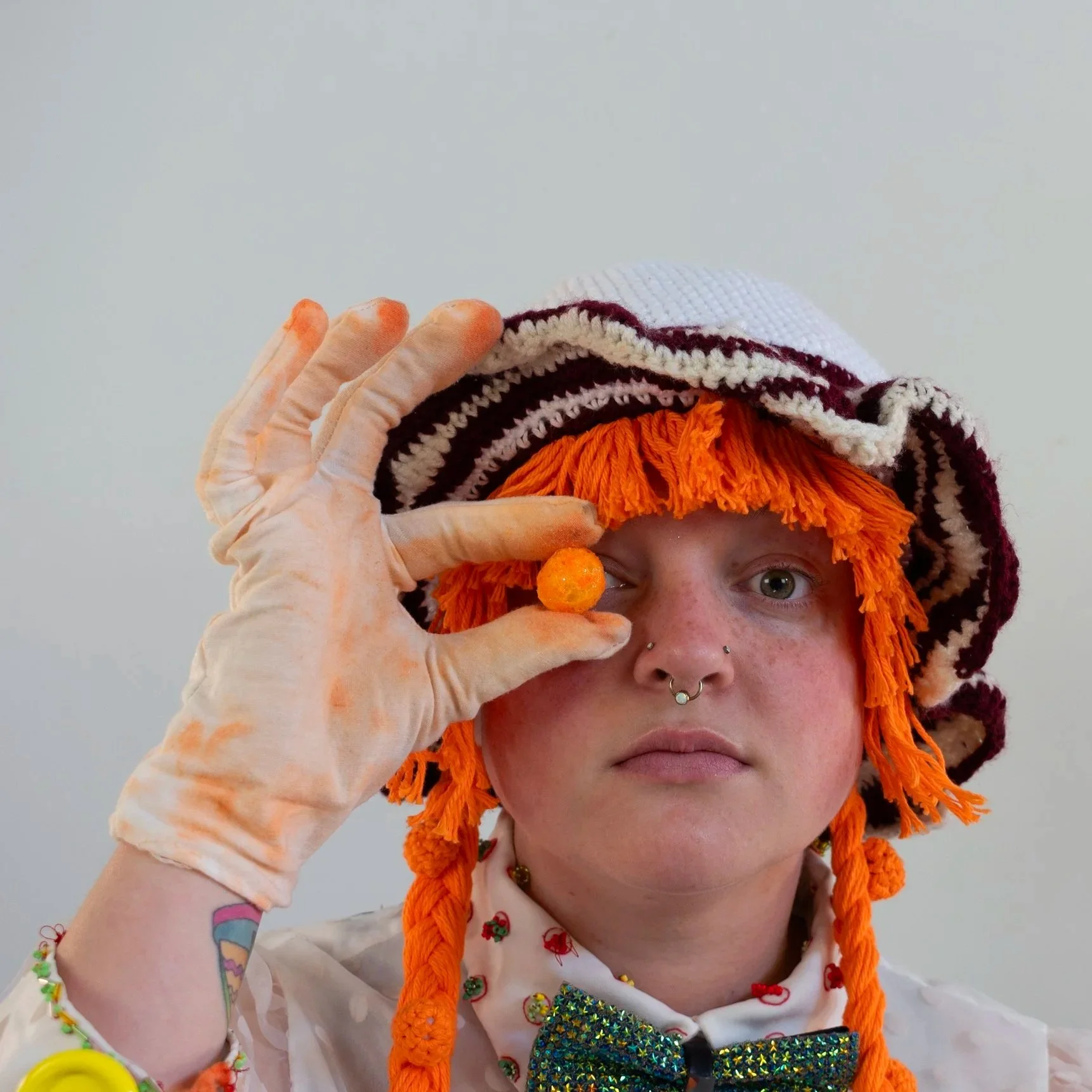 Bea is a lesbian white women with short light brown hair. She is wearing a crocheted clown hat and white gloves, while holding a cheese ball snack up to her right eye.