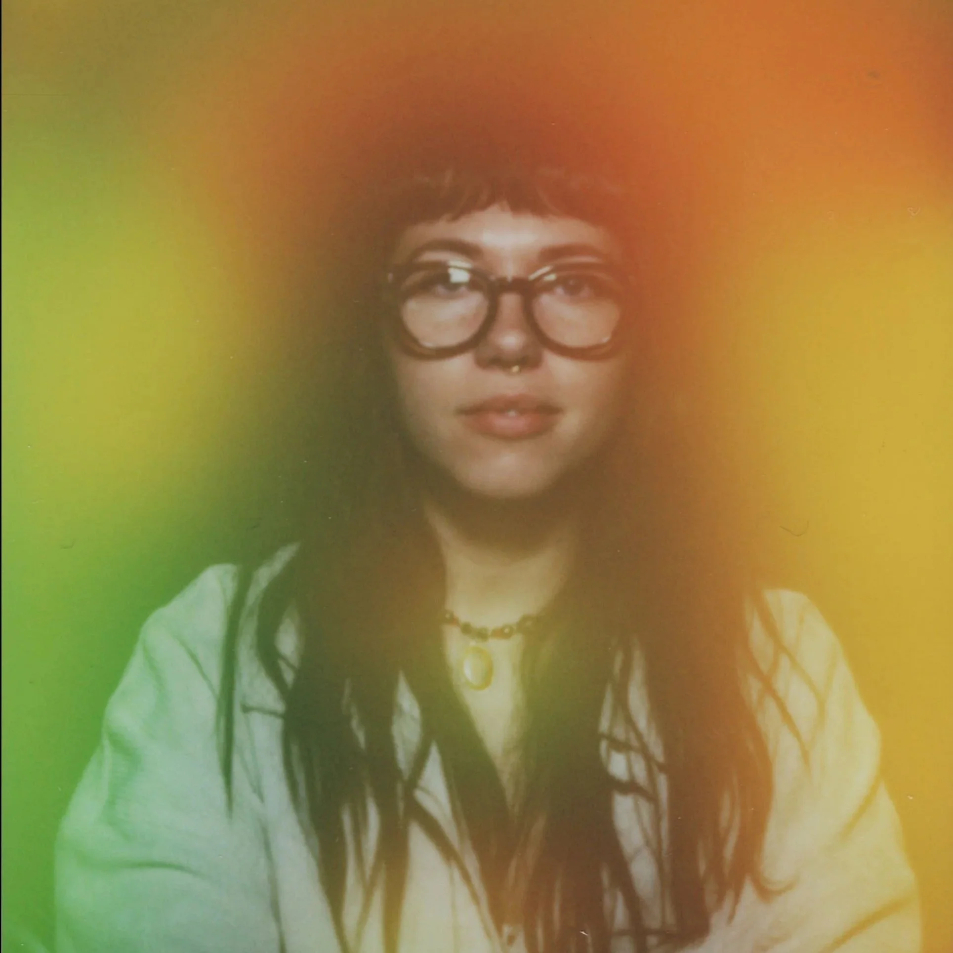 Bri is a bisexual white woman with long dark hair. Bri is wearing brown rimmed glasses and a white button up. The image is an aura photo featuring green, yellow and red around Bri’s head.